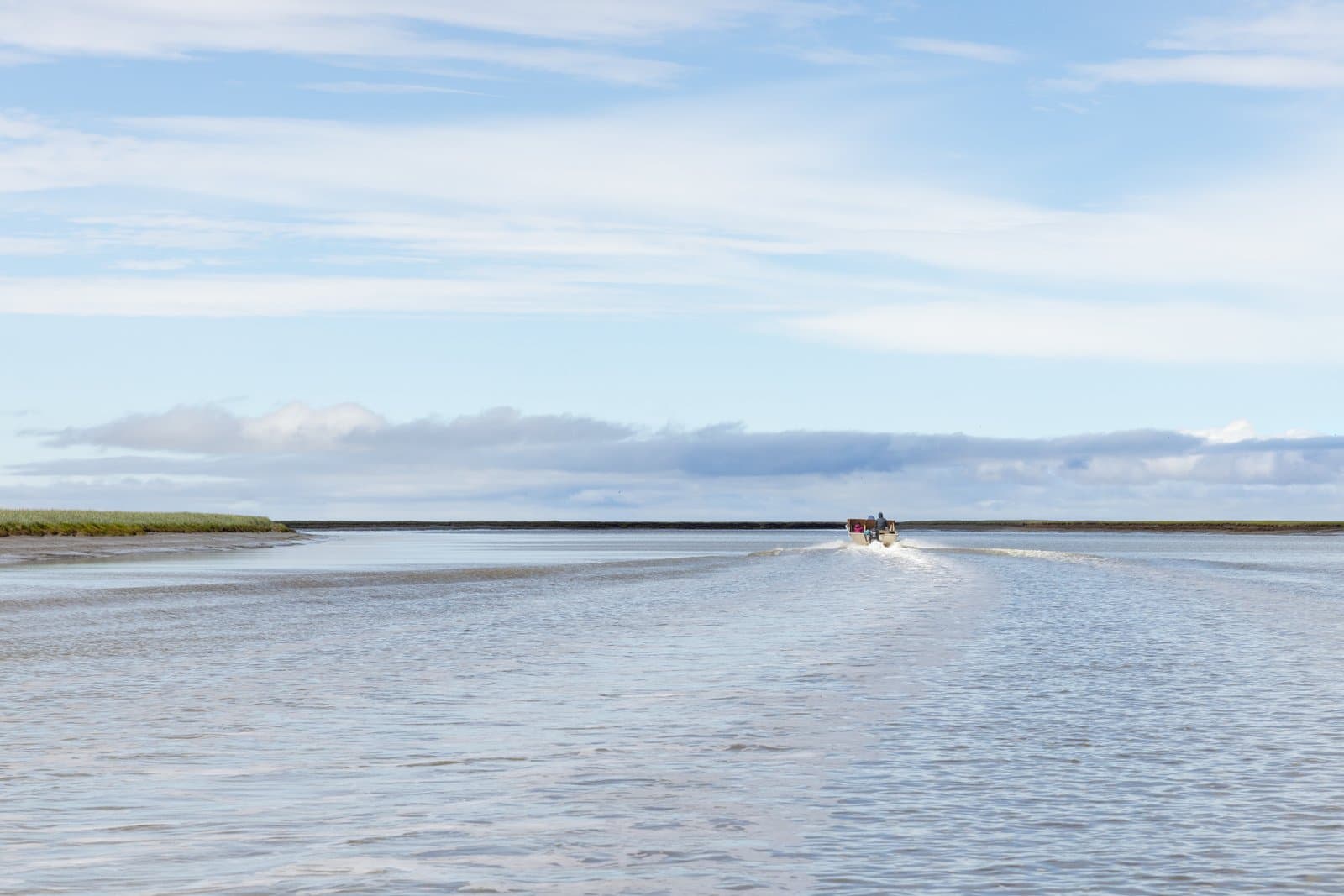 A small skiff travels along the calm waters of the Ningliqvak River under a bright, cloudy sky in rural Alaska.