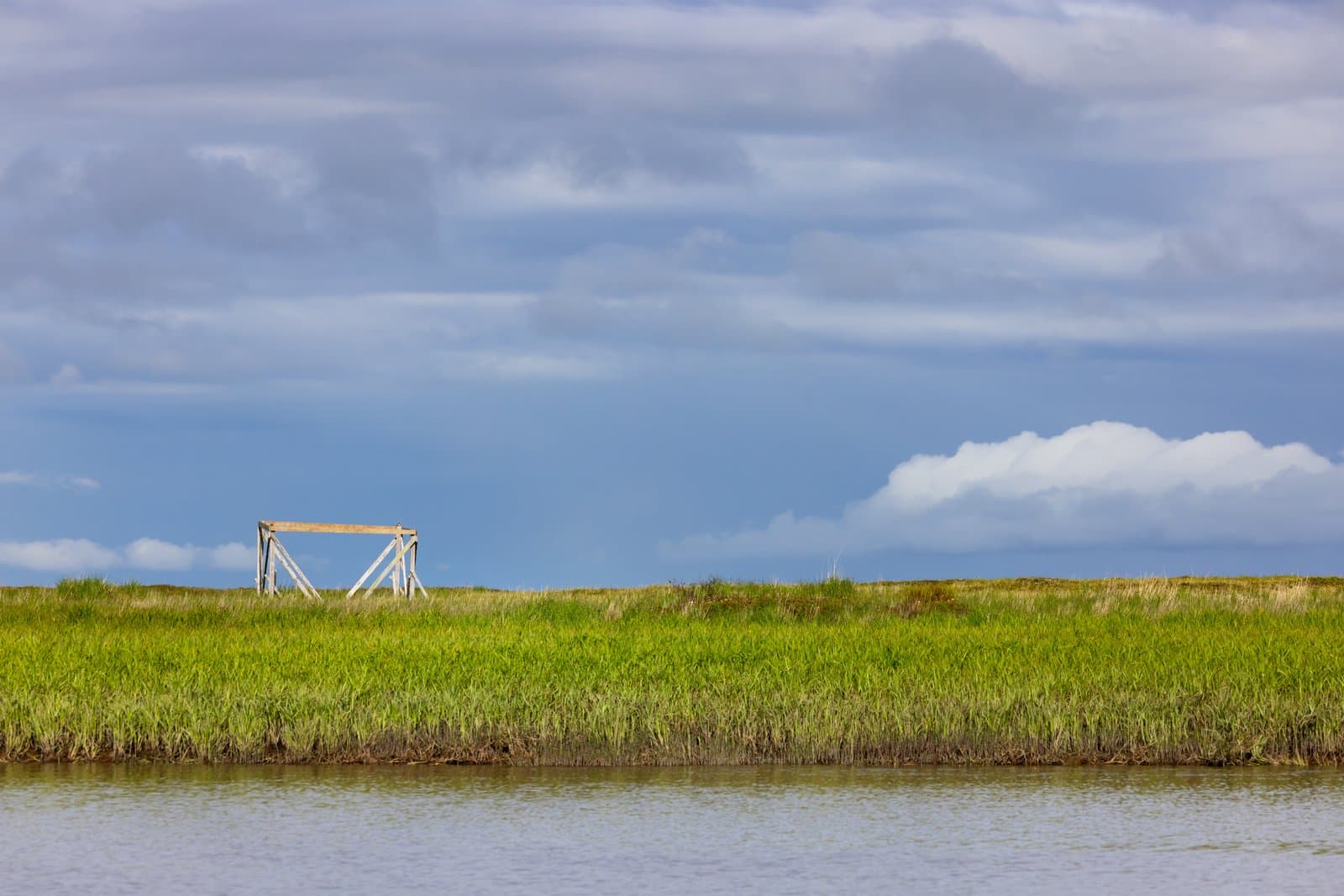 A wooden fish drying rack stands in a grassy field near a body of water under a cloudy sky in Alaska.