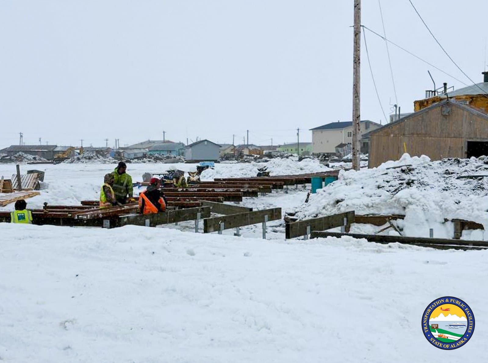 Construction workers in high-visibility gear assemble a wooden structure on a snow-covered site in the community of Kipnuk, Alaska.