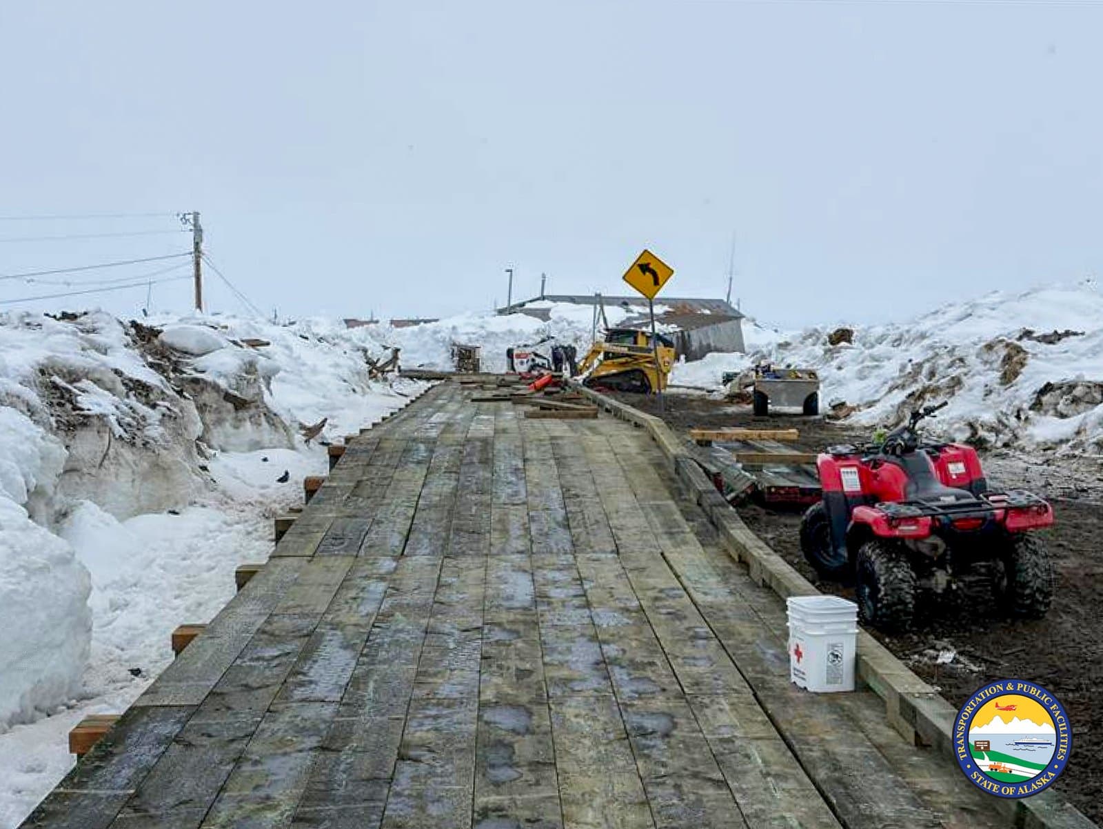 A wooden boardwalk under construction in Kipnuk, Alaska, features heavy machinery, a red ATV, and surrounding snowbanks during the spring season.