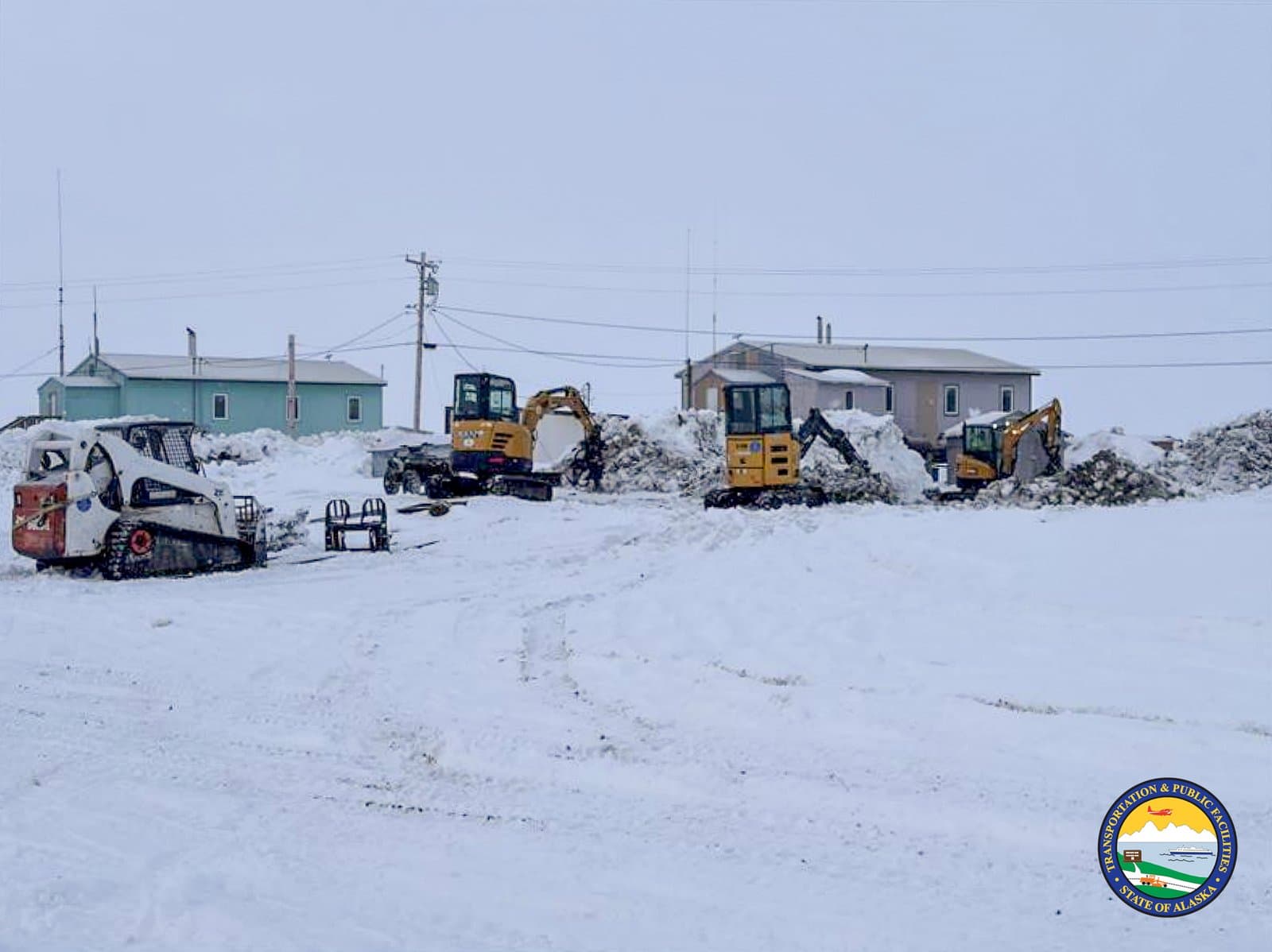Yellow excavators and a white skid steer clear deep snow piles near residential buildings in the community of Kipnuk, Alaska.
