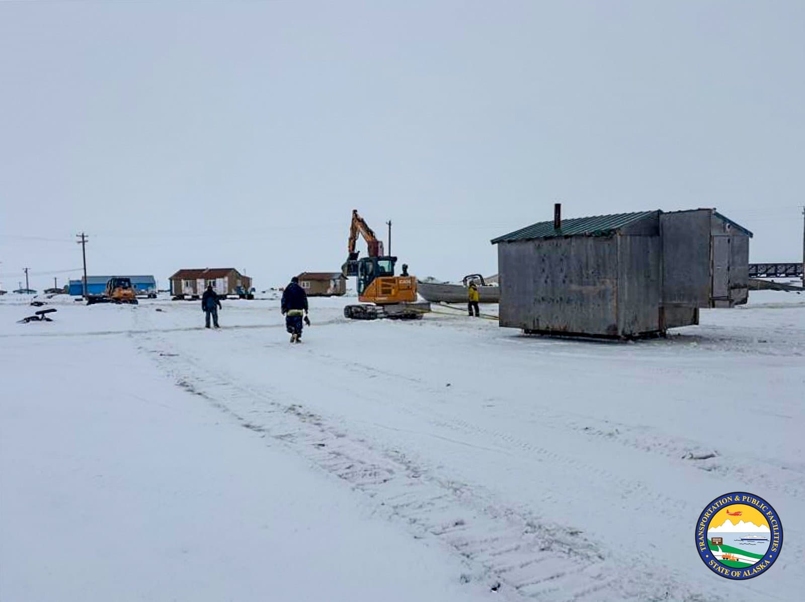 Workers use a yellow excavator to relocate a small wooden structure across a snow-covered landscape in the village of Kipnuk, Alaska.