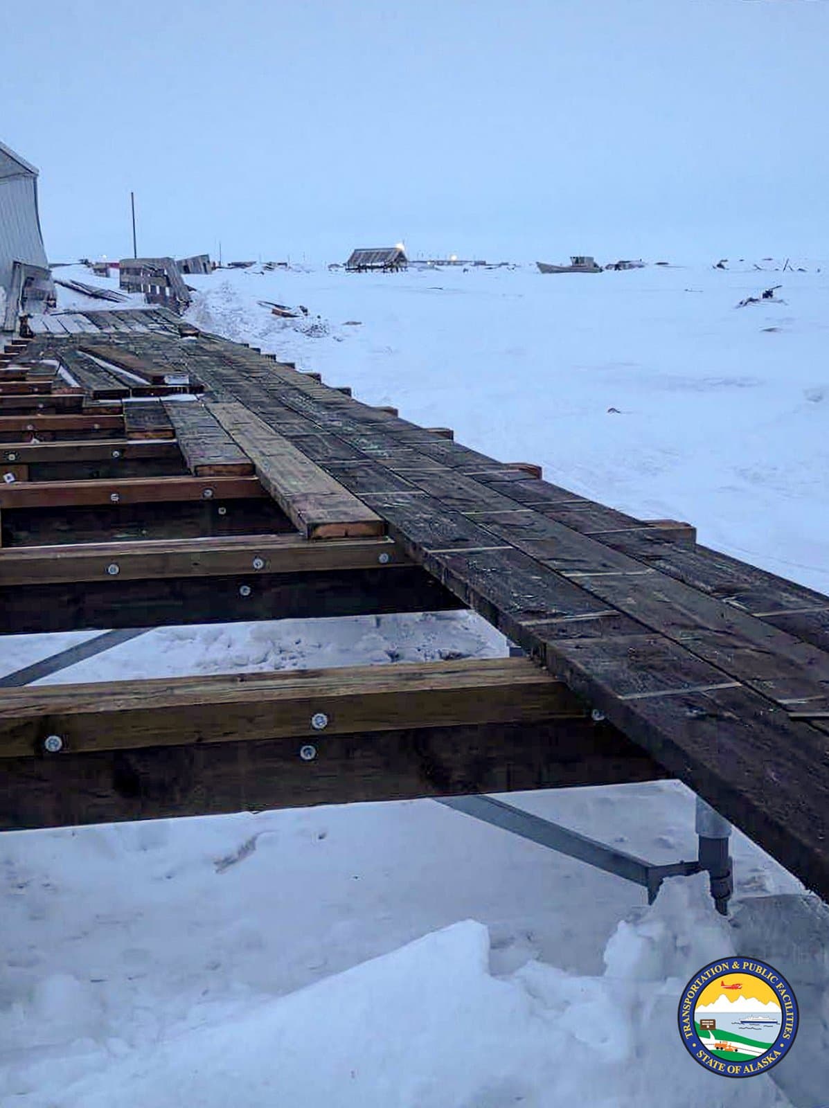 A wooden boardwalk under construction stretches across a snow-covered landscape in the rural community of Kwigillingok, Alaska, during the spring season.