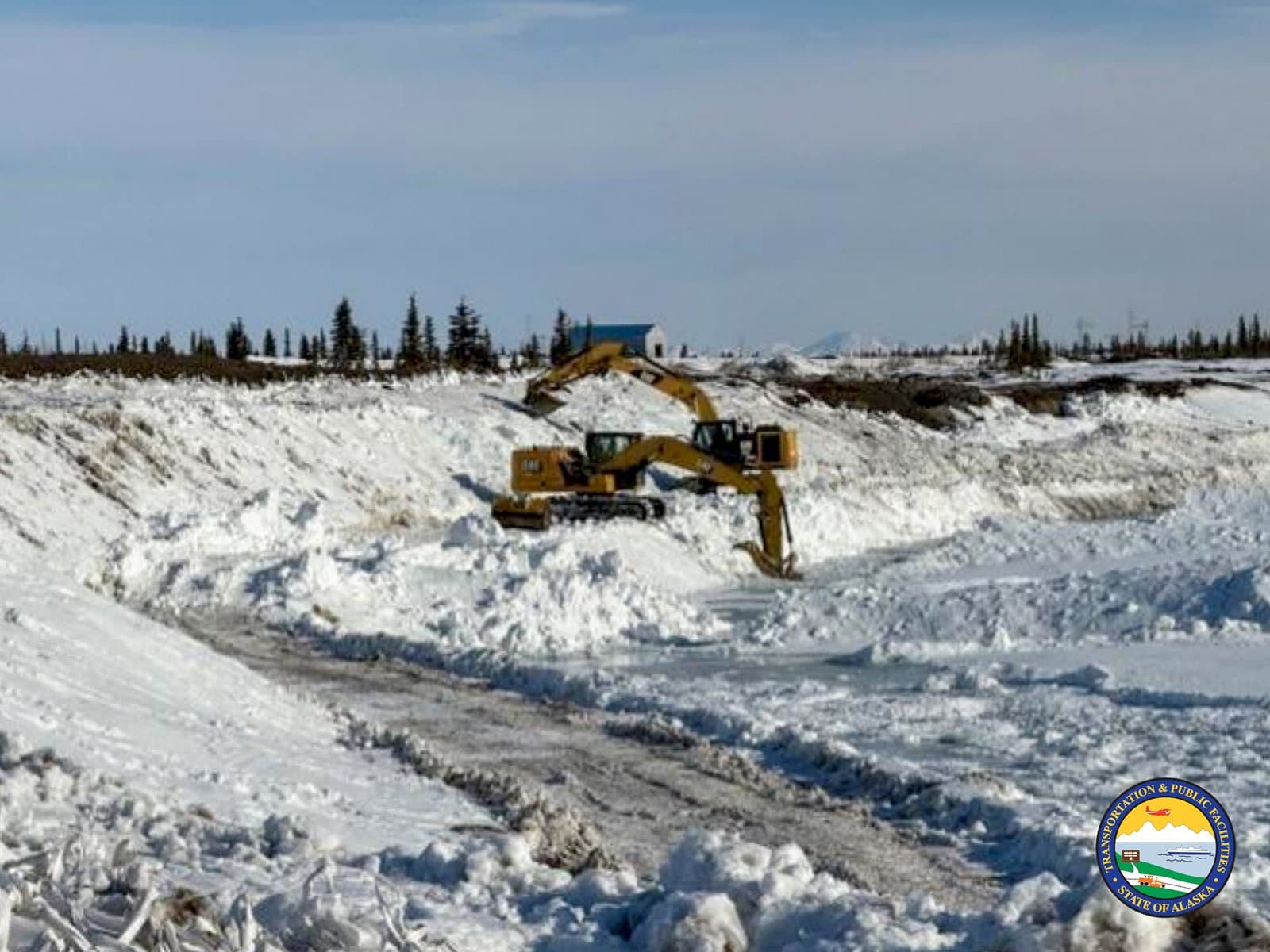 A yellow excavator clears deep snow from a road or site in Noatak, Alaska, during a spring maintenance operation.