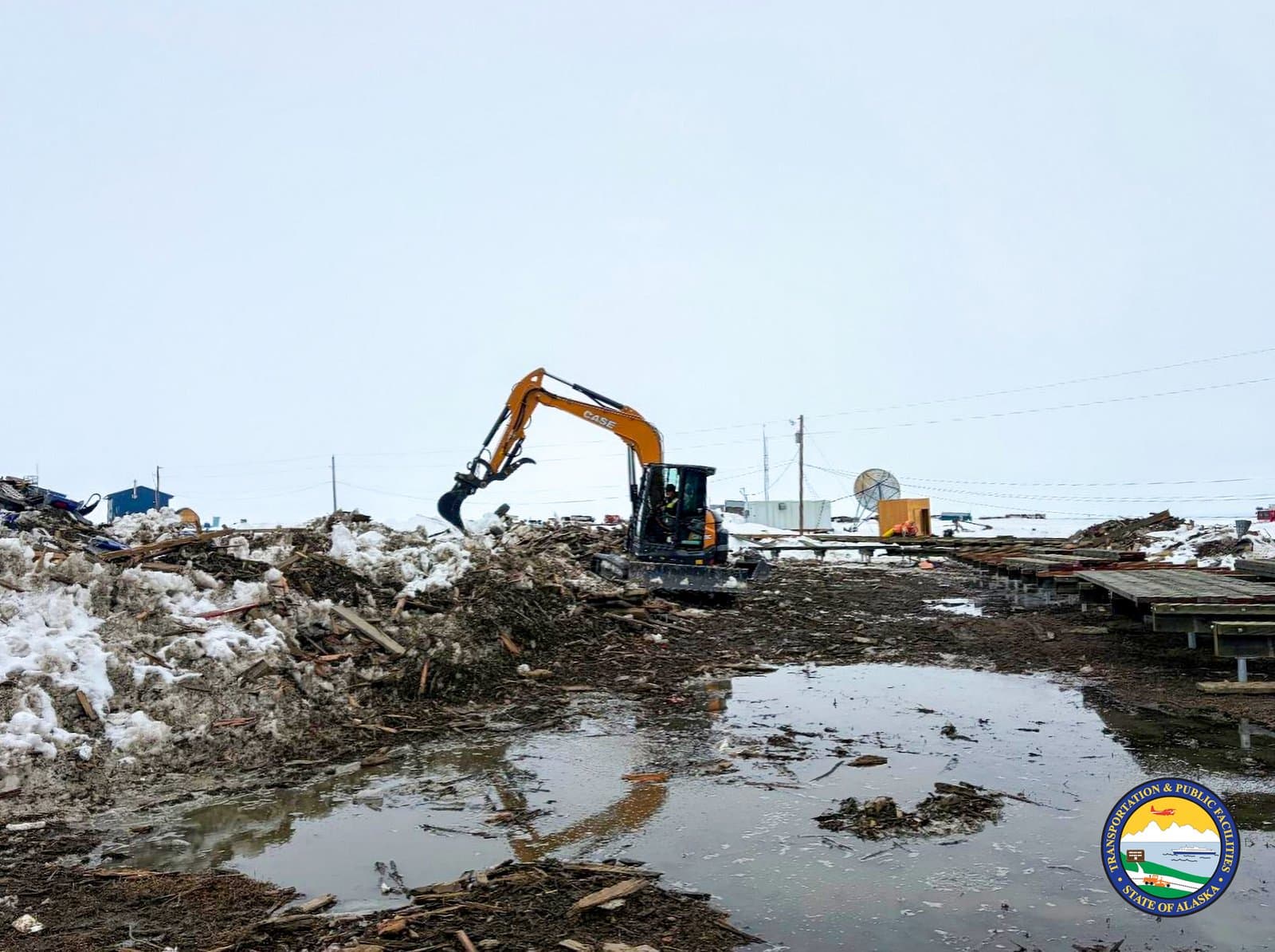 An orange Case excavator works to clear debris and melting snow from a muddy, water-logged site in the community of Kipnuk, Alaska.
