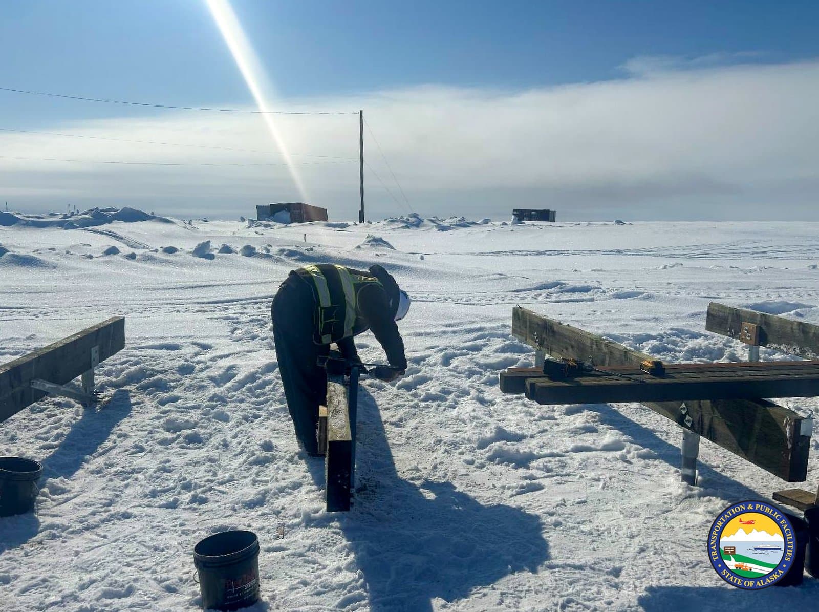 A construction worker in high-visibility gear installs wooden beams on a snowy, flat landscape in Kwigillingok, Alaska, during early April.