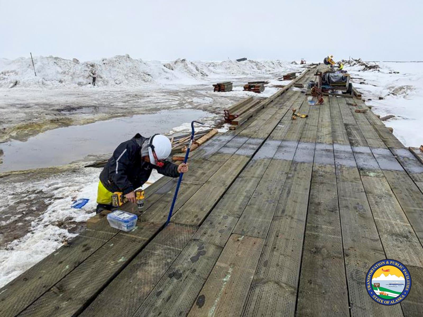 A construction worker uses a drill and pry bar to repair a wooden boardwalk in the snowy, coastal community of Kipnuk, Alaska.