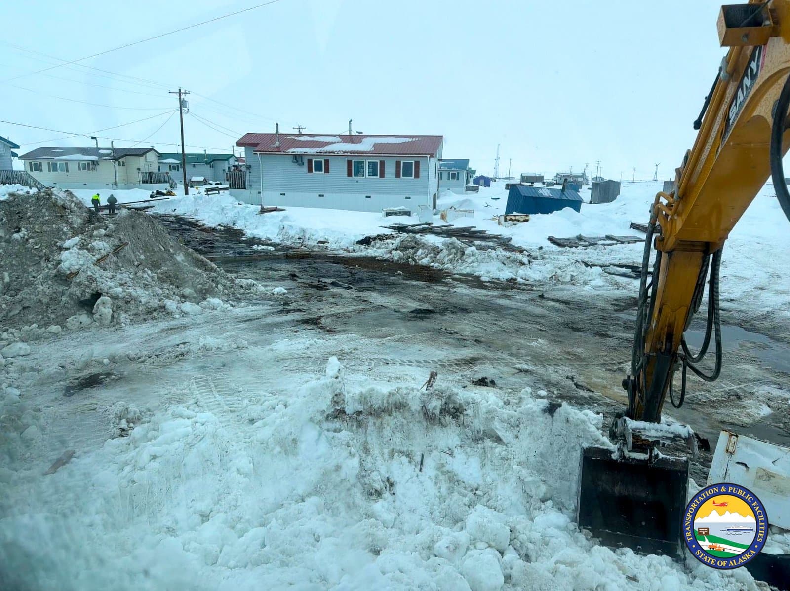 A yellow excavator clears snow and debris from a muddy, slushy road in the community of Kwigillingok during early April.