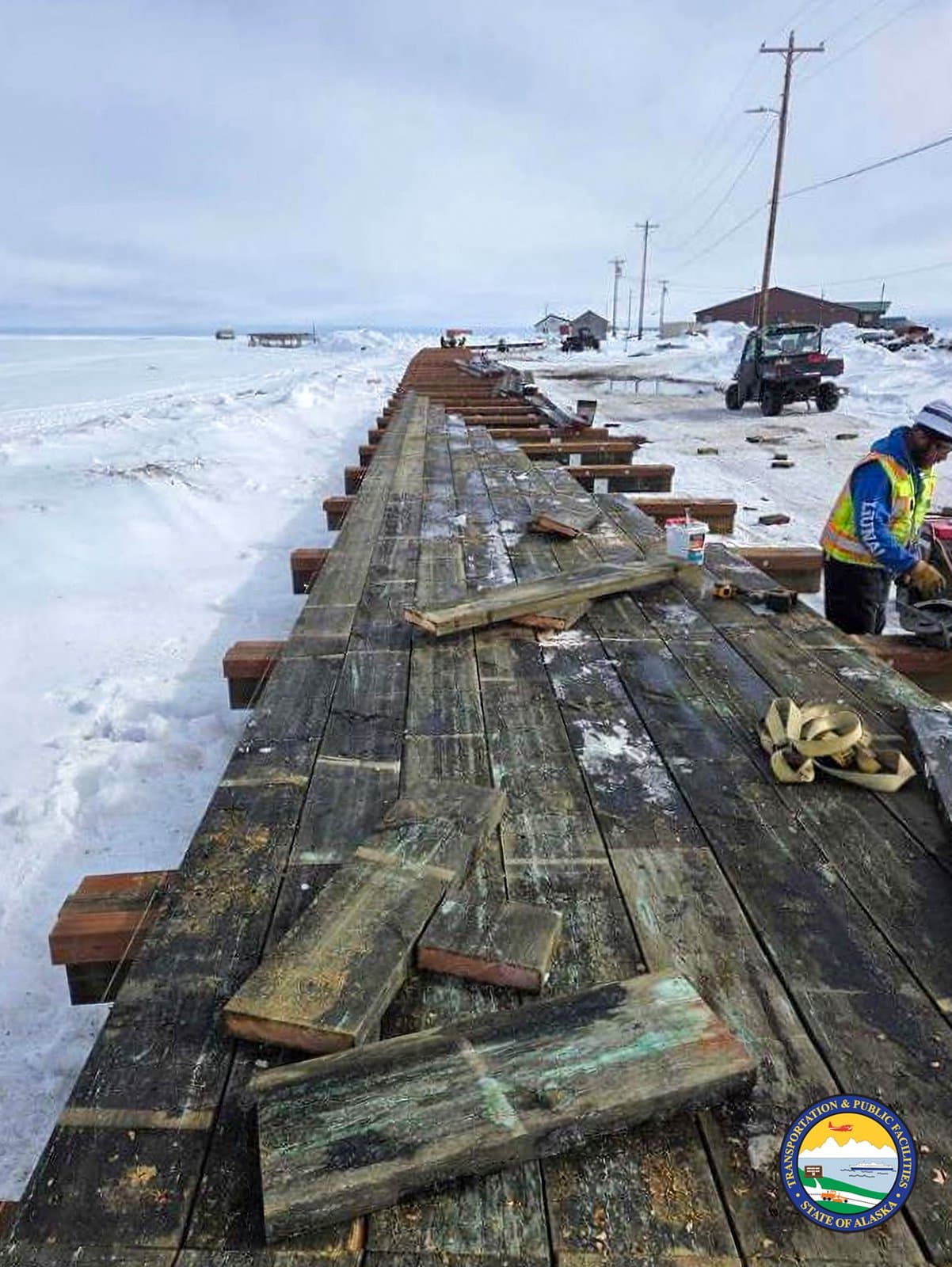 A worker installs wooden planks on a boardwalk in Kwigillingok, Alaska, surrounded by snow-covered terrain under a cloudy spring sky.