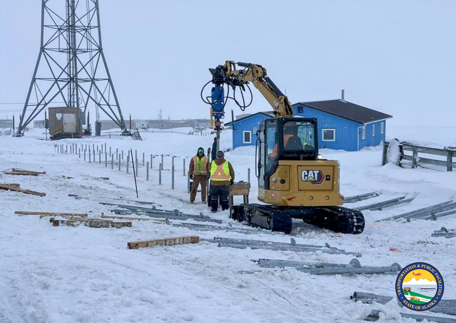 Construction workers operate a Caterpillar excavator to install metal posts into the frozen, snow-covered ground in the community of Kipnuk, Alaska.