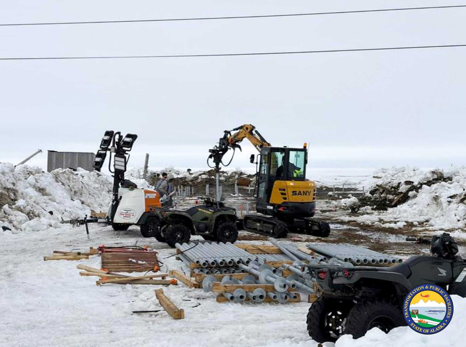A Sany excavator and all-terrain vehicles operate on a snowy construction site in Kipnuk, Alaska, surrounded by piles of metal piping.