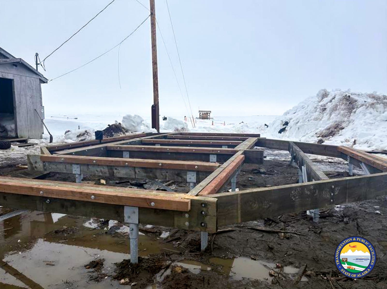 A wooden foundation frame sits on metal pilings in the muddy, snow-covered landscape of Kwigillingok during the spring thaw.