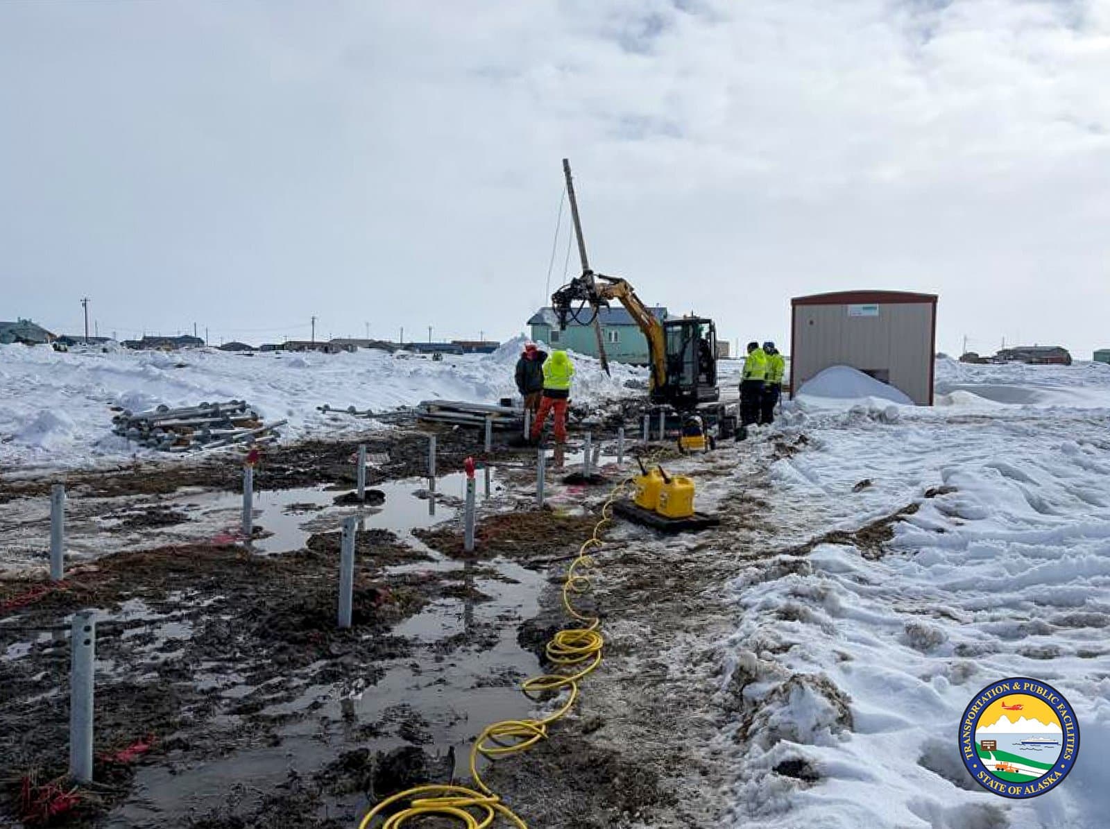 Construction workers operate a small excavator to install metal foundation pilings in the muddy, snow-covered ground of Kipnuk, Alaska.