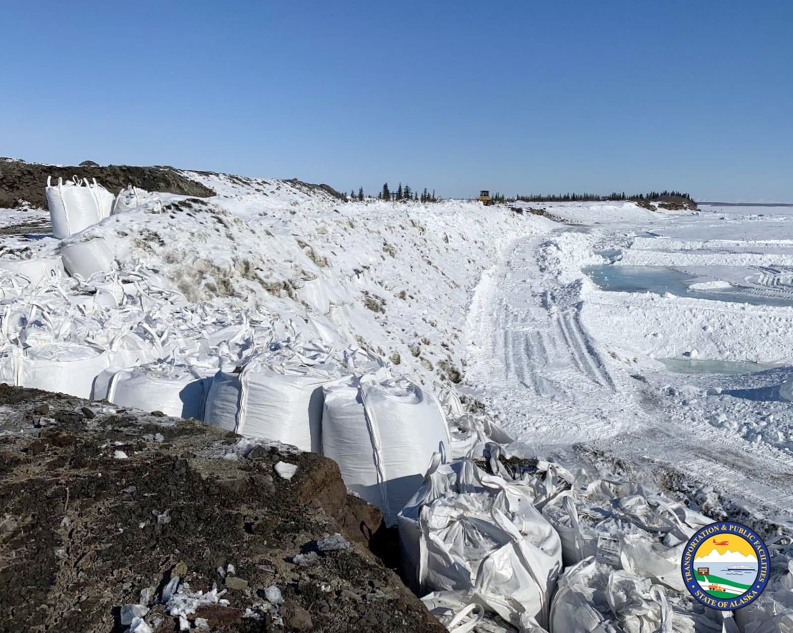 Large white sandbags line a snow-covered embankment near the frozen Noatak River, serving as flood protection infrastructure in rural Alaska.