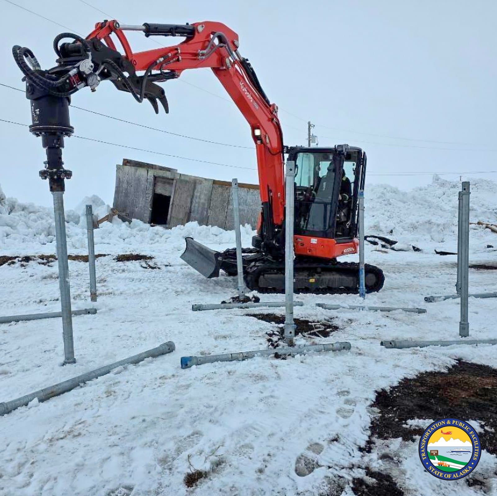 A Kubota excavator equipped with a pile driver attachment installs metal posts into the frozen, snow-covered ground in Kwigillingok, Alaska.