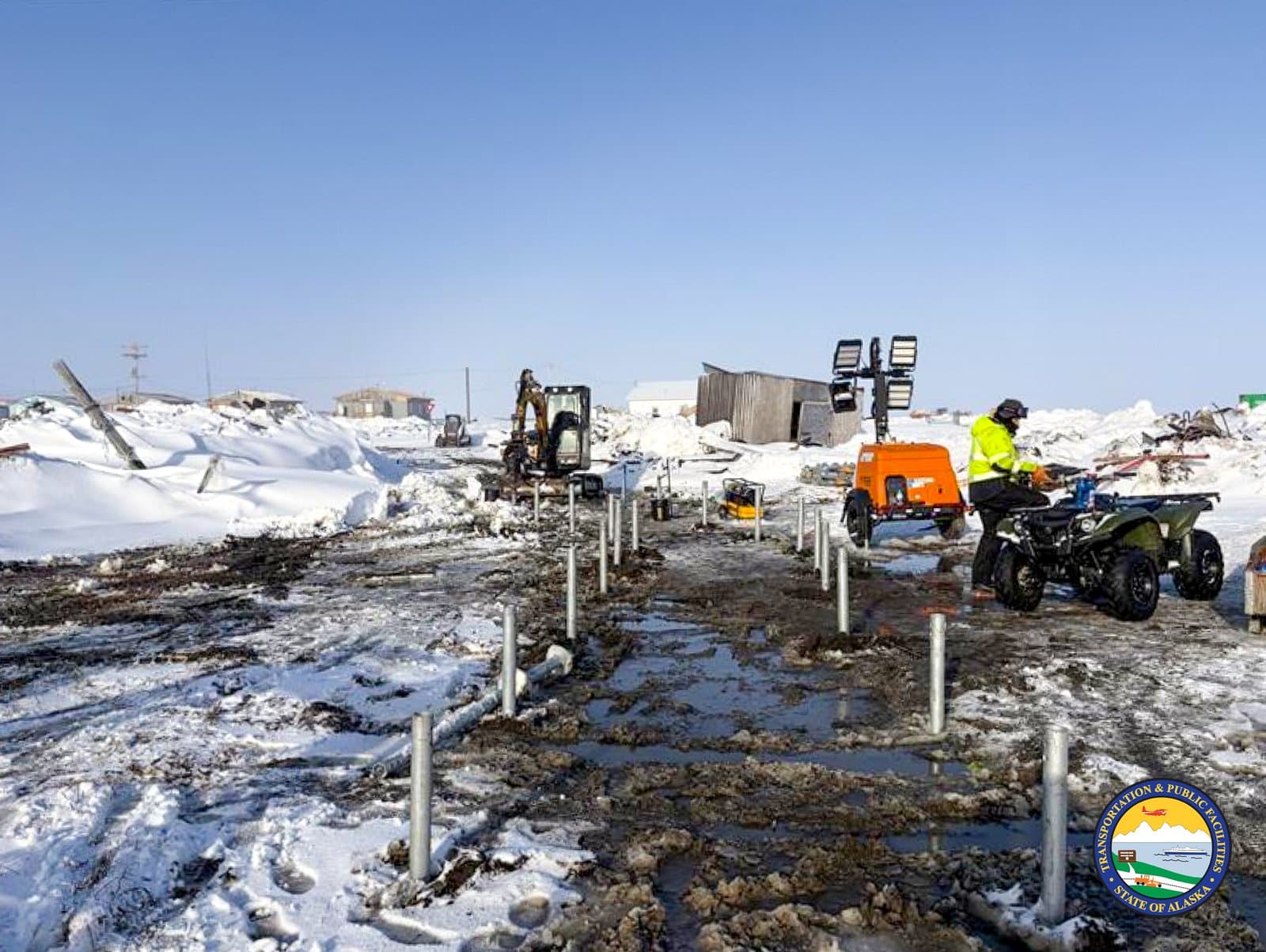 A worker on an ATV monitors construction equipment and metal posts installed in the muddy, snow-covered ground of Kipnuk, Alaska.
