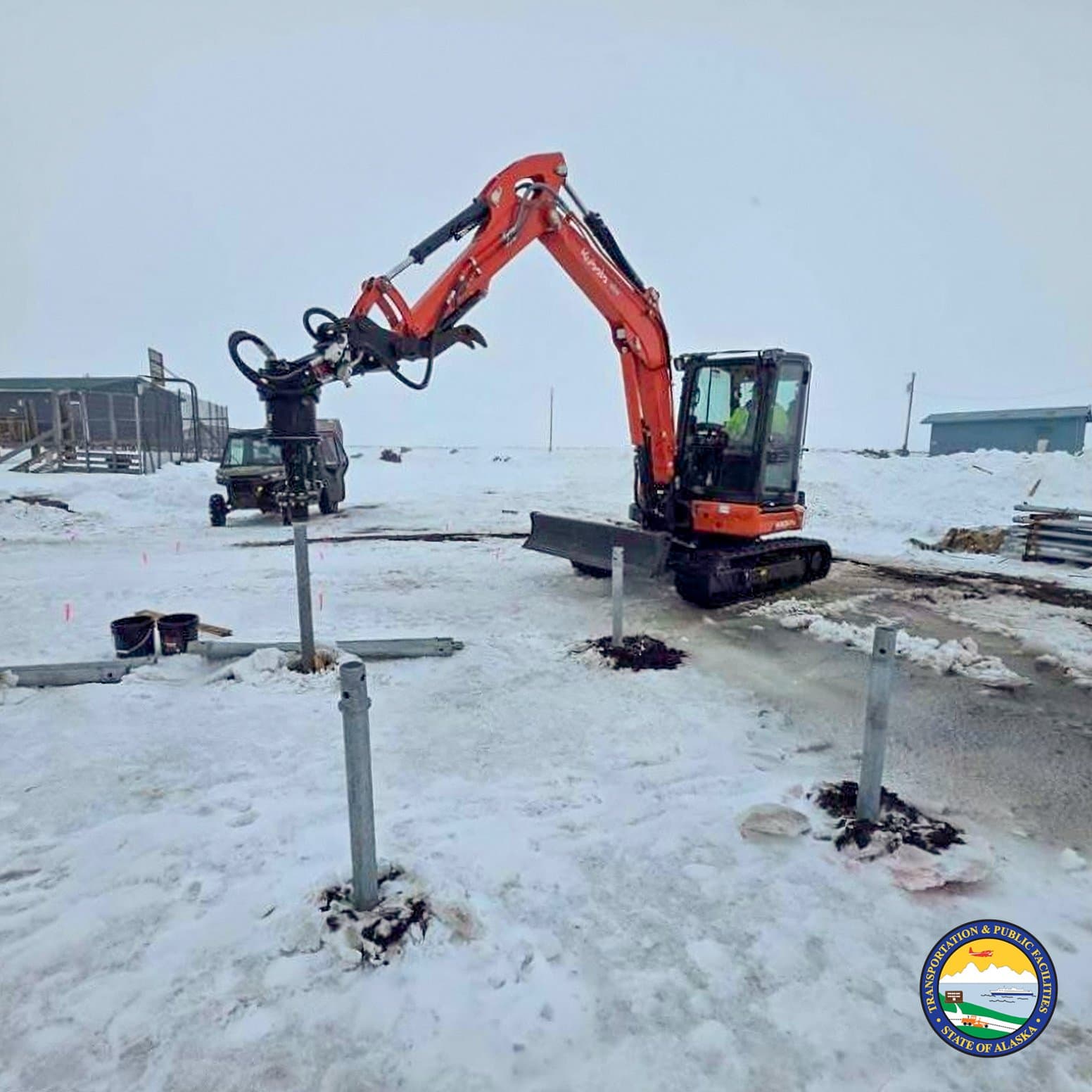 An orange excavator with a hydraulic attachment installs metal posts into the frozen, snow-covered ground in the community of Kwigillingok, Alaska.