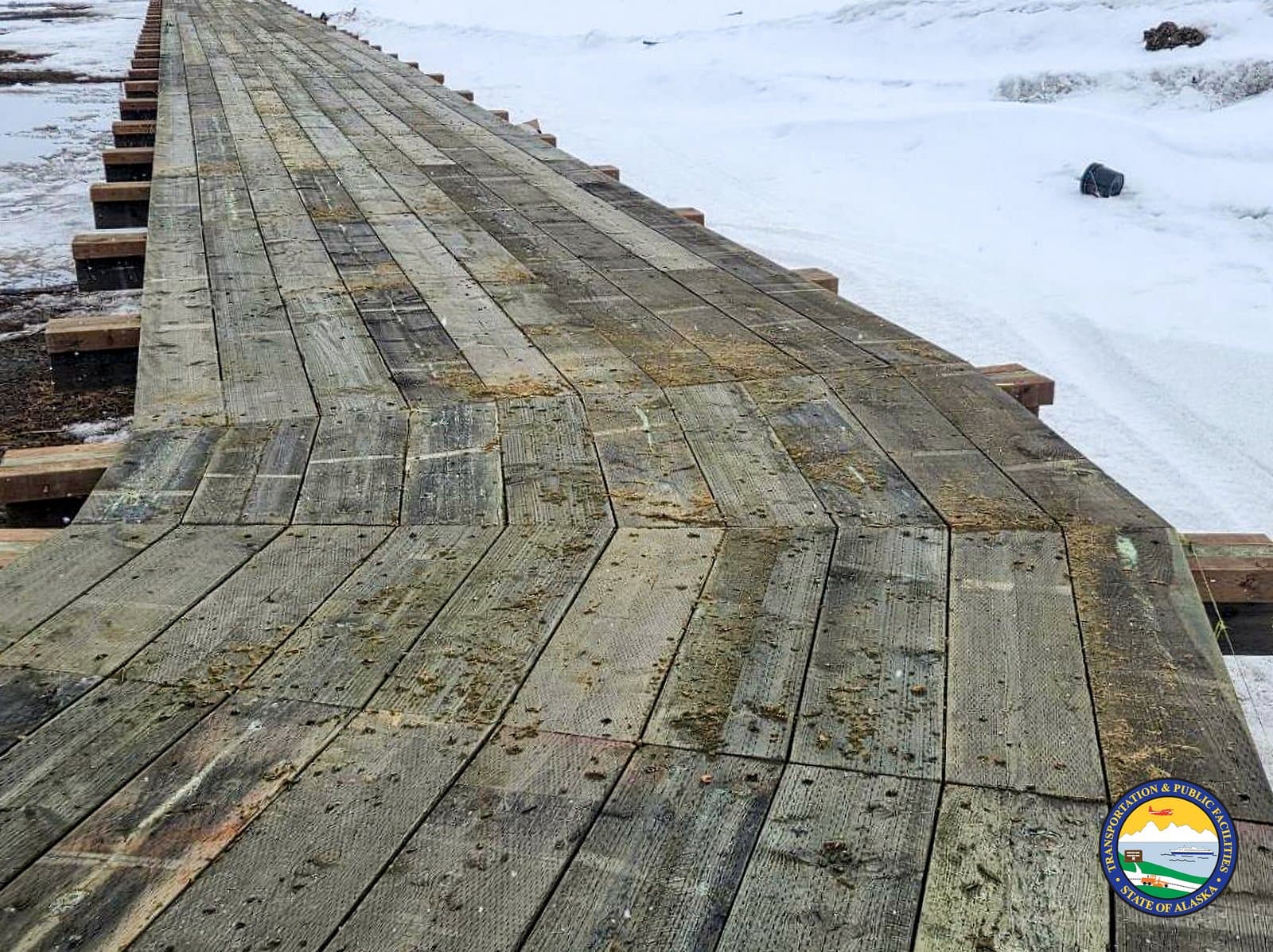 A weathered wooden boardwalk stretches across a snow-covered landscape in Kwigillingok, Alaska, during the spring season.