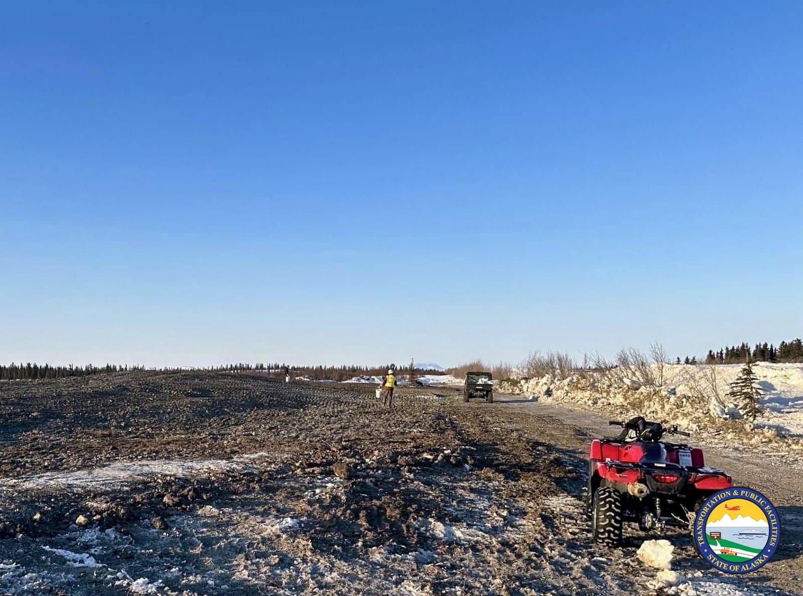 A worker in a high-visibility vest walks across a muddy, partially snow-covered airstrip in Noatak, Alaska, while a red ATV sits nearby.