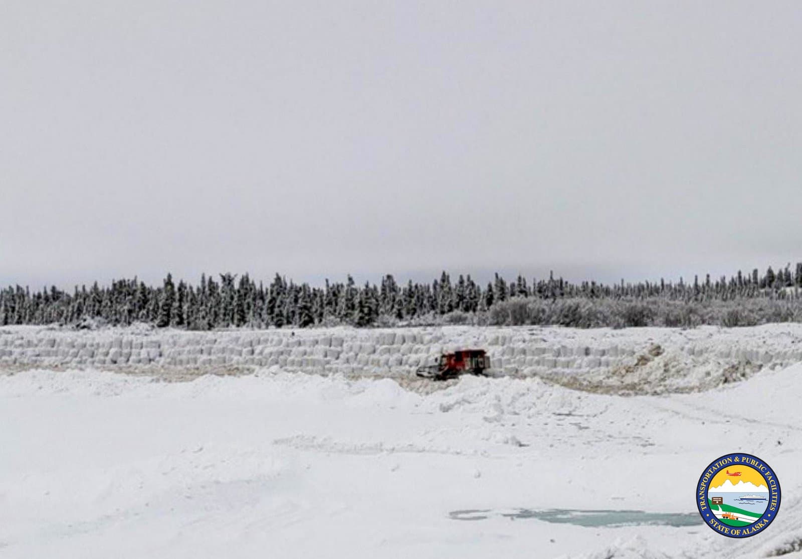 A red snow removal vehicle clears a path through deep snow near a line of white sandbags in Noatak, Alaska.
