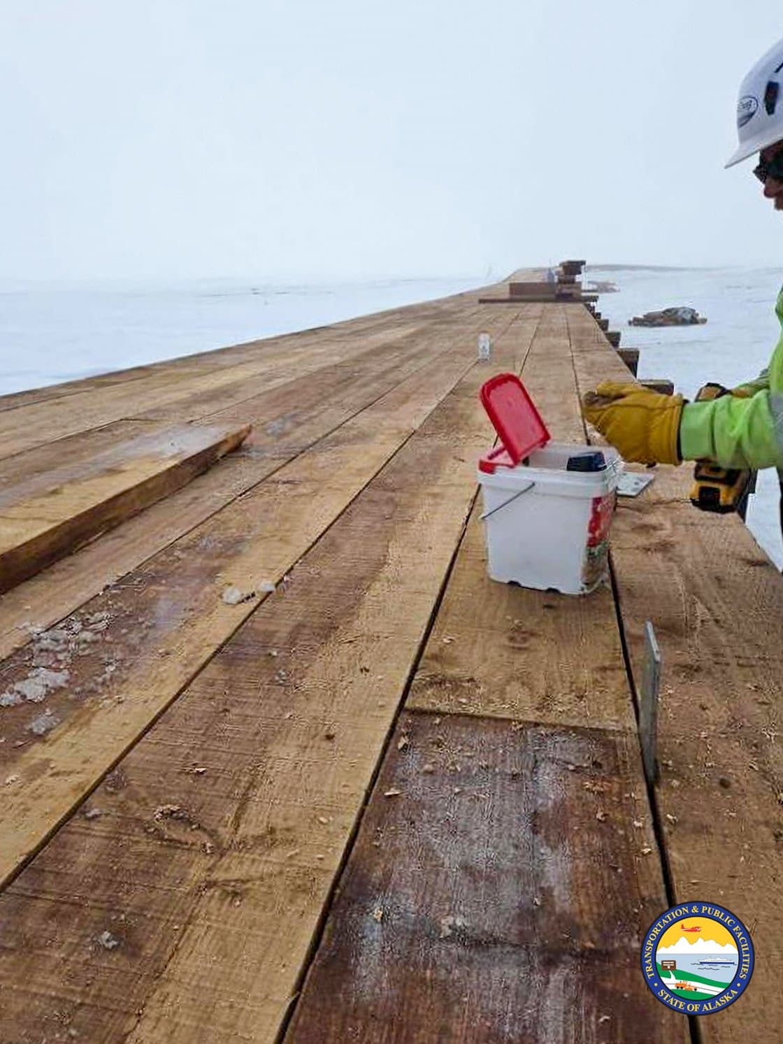 A construction worker in high-visibility gear works on a wooden boardwalk in Kwigillingok, Alaska, during the spring season.
