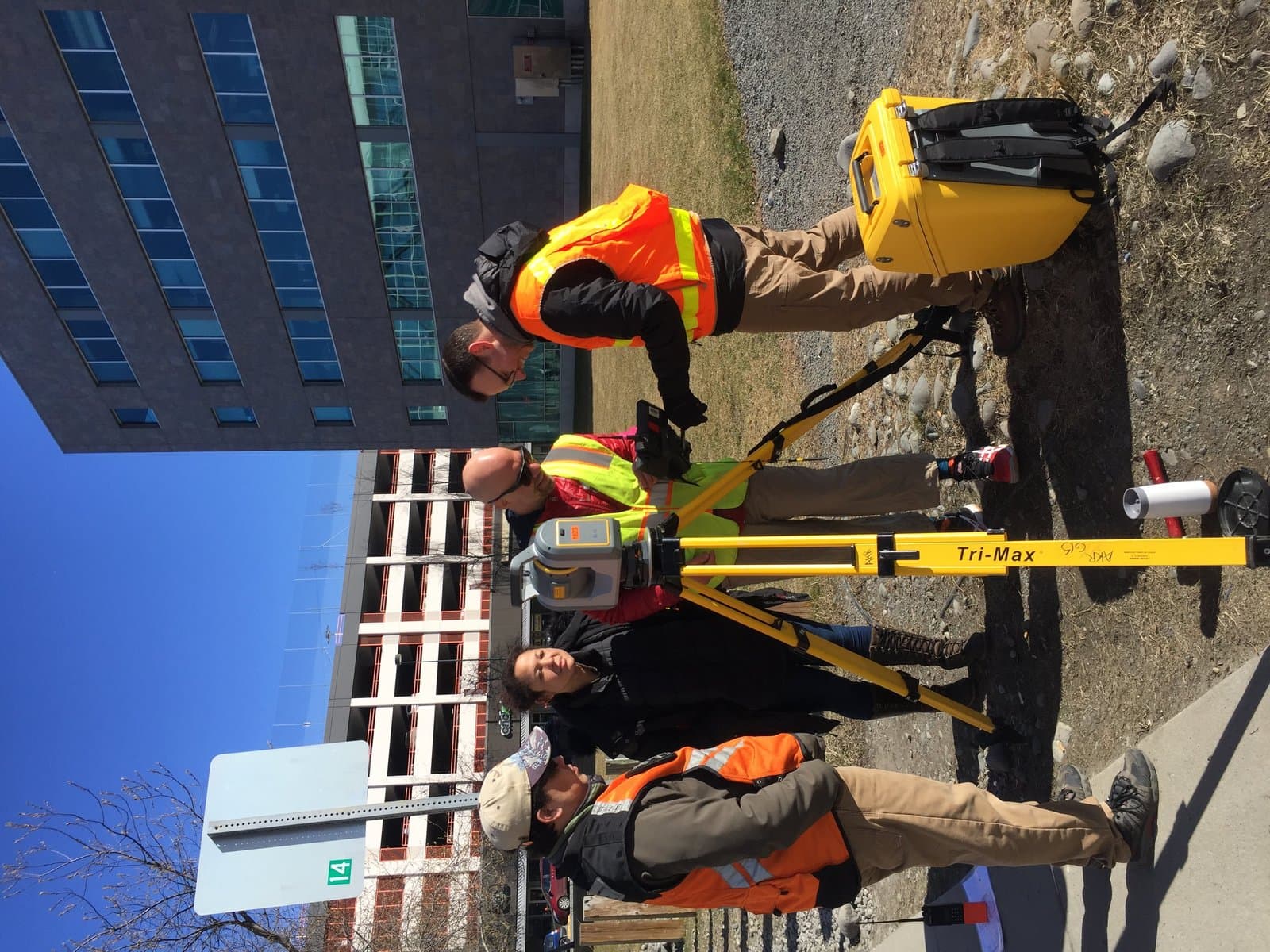 Four individuals, some wearing safety vests, are conducting a land survey outdoors with specialized equipment, including a total station on a tripod and a yellow case. Modern office buildings and a parking garage are visible in the background under a clear blue sky.