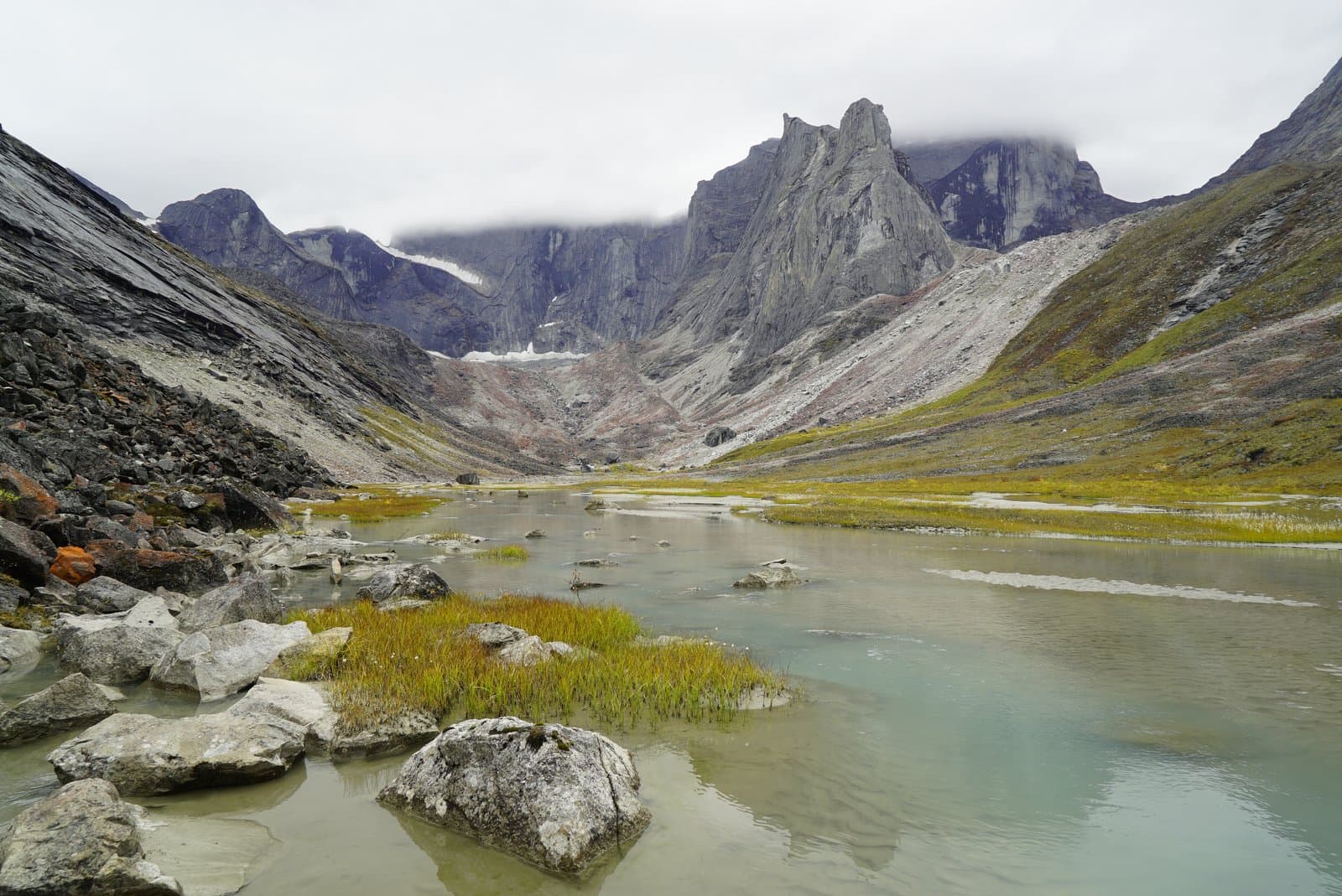 A wide shot of a glacial lake in a mountainous valley under a cloudy sky. Jagged, rocky peaks rise on both sides, with a glacier visible in the distance. The lake's edges are lined with green and yellow grasses and large rocks.