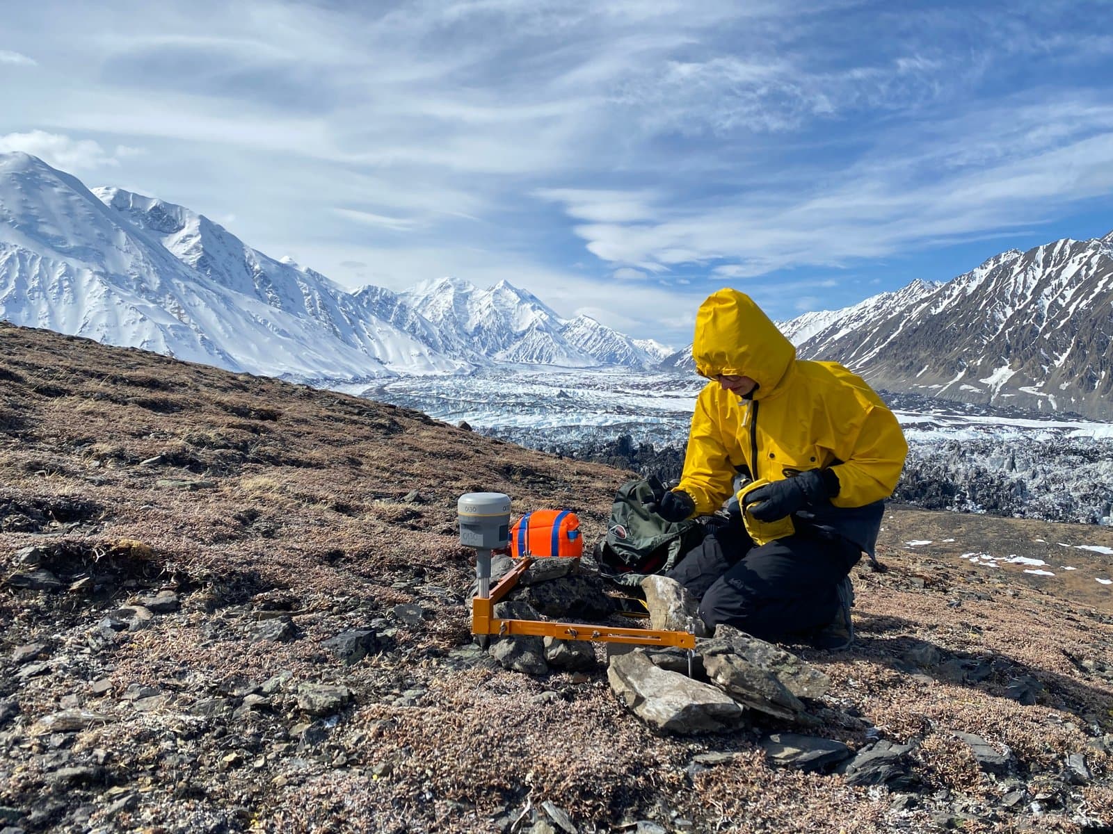 A person in a bright yellow hooded jacket kneels on rocky, sparse ground, adjusting scientific equipment next to a backpack. In the background, a vast glacier winds through towering, snow-capped mountains under a partly cloudy sky.