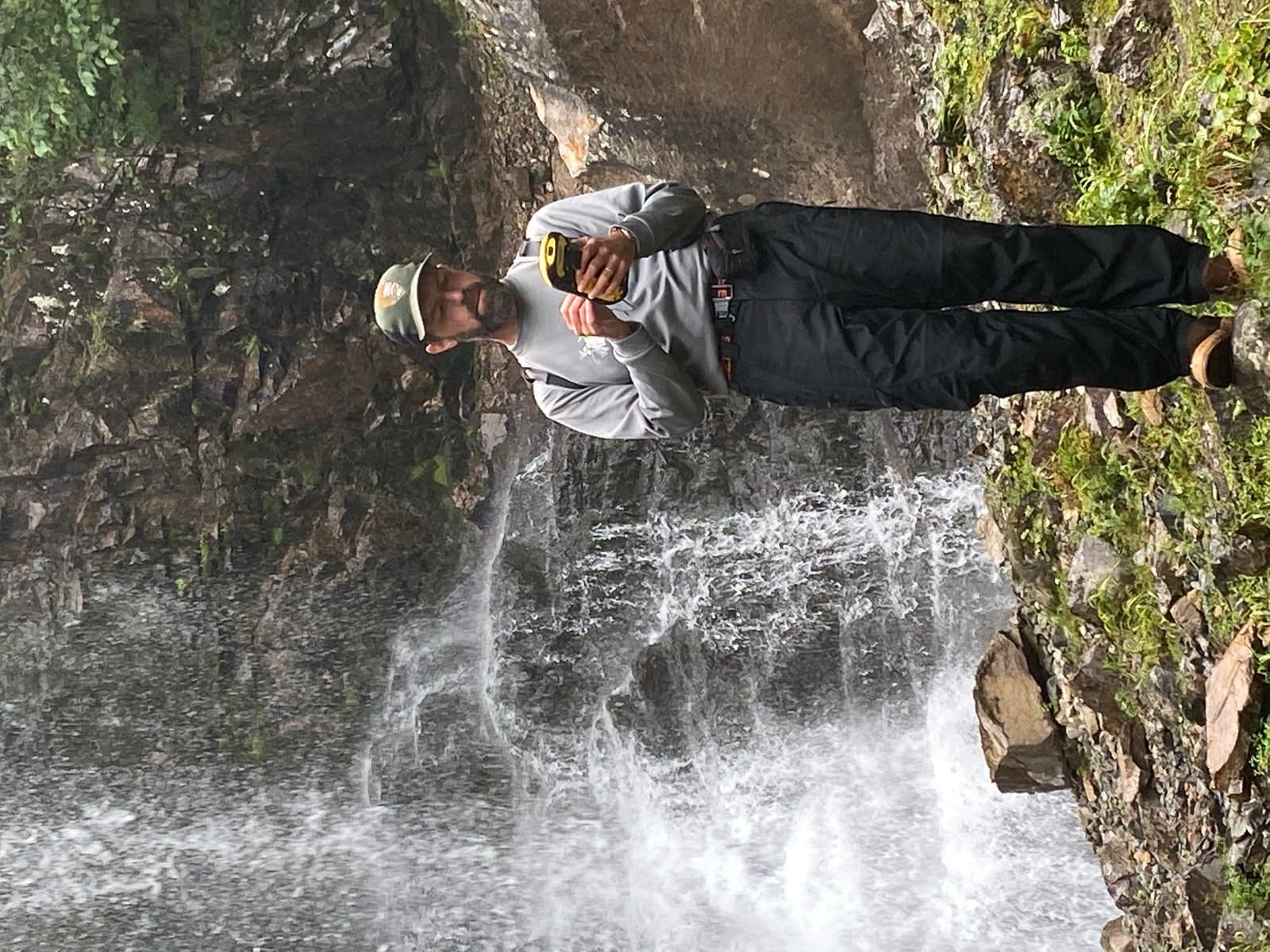 A man in outdoor gear, wearing a cap and holding a yellow device, stands beside a powerful waterfall, seemingly taking a photo. The background features rocky cliffs and lush green vegetation, with water cascading down into a turbulent pool.