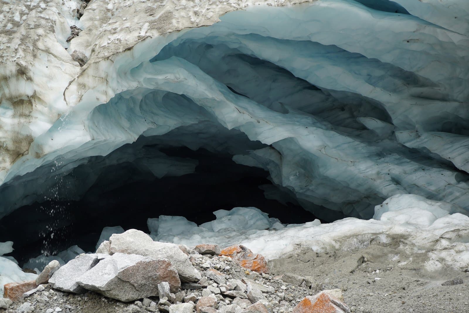 A detailed view of the entrance to an ice cave in Arrigetch Valley, showcasing intricate layers of translucent blue ice forming wavy patterns. Water drips from the cave ceiling, and a scattering of rocks and gravel lies at the opening.