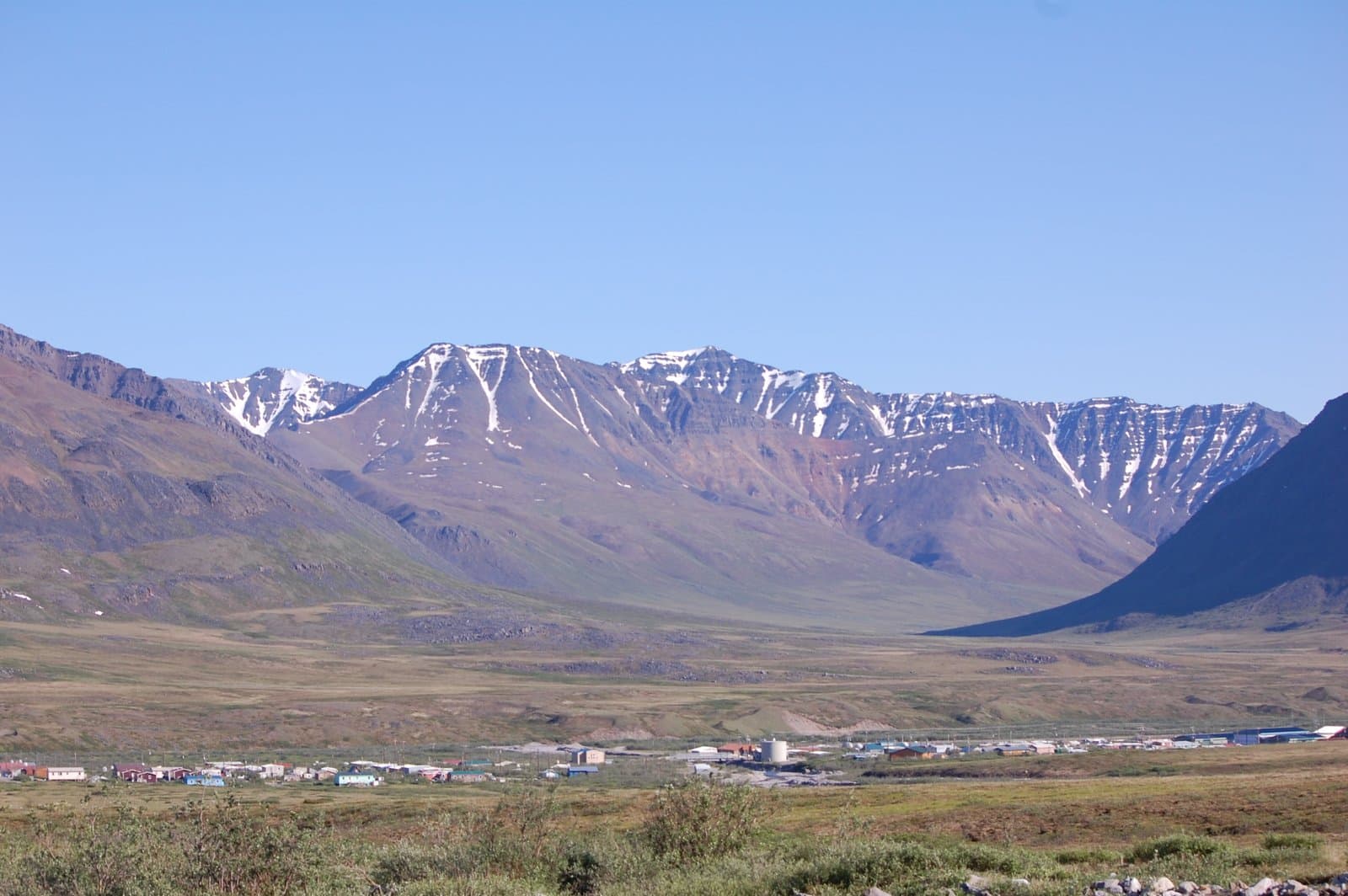 A wide shot of Anaktuvuk Pass, an Alaskan village, nestled in a vast valley with snow-capped mountains of the Brooks Range in the background under a clear blue sky. The foreground features green and brown tundra.