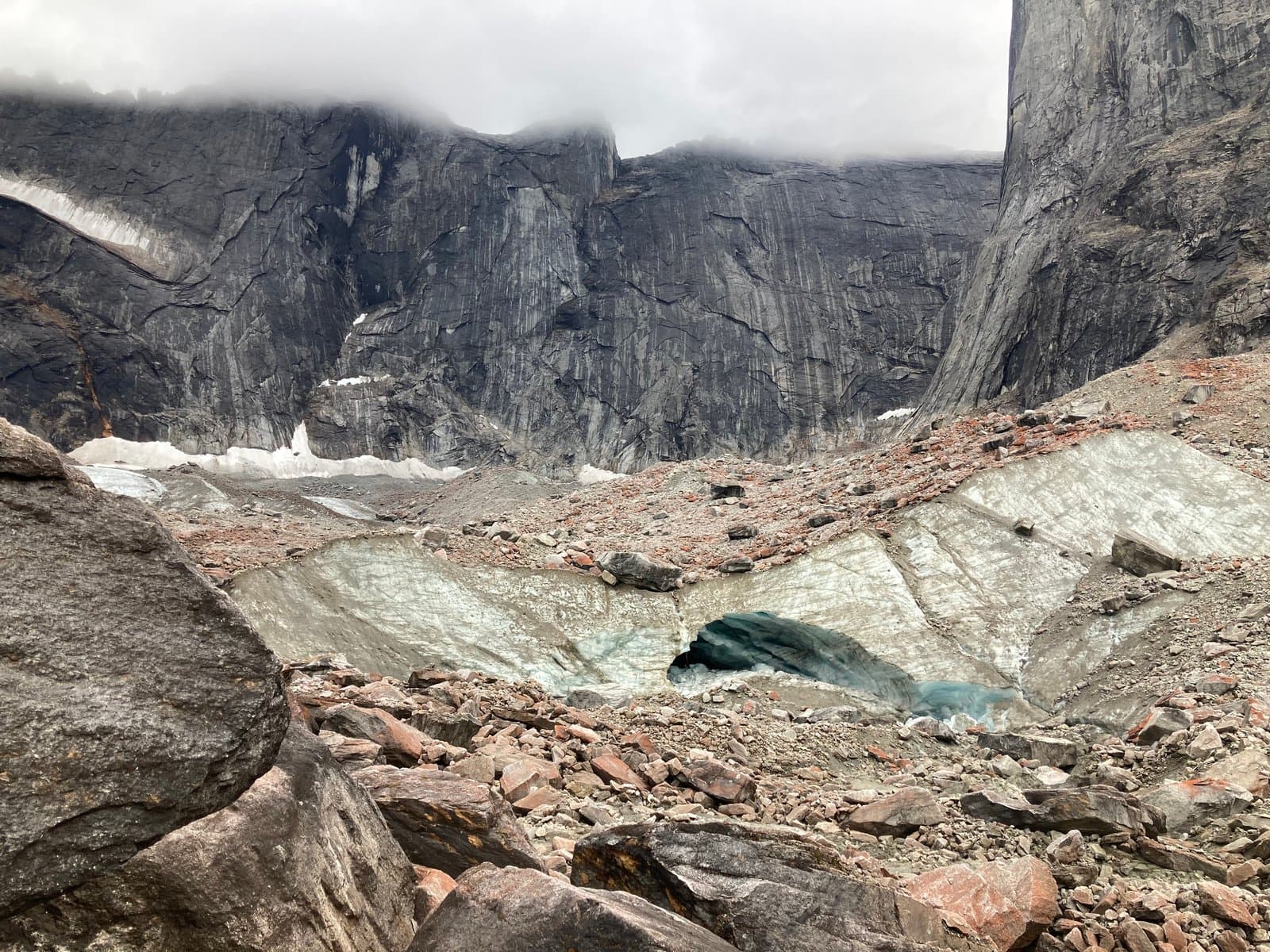 A large, dark grey mountain range rises under a cloudy sky, with a glacier at its base. The glacier is covered in rocks and debris, and features a prominent, deep blue ice cave in the center, surrounded by more rocky terrain in the foreground.