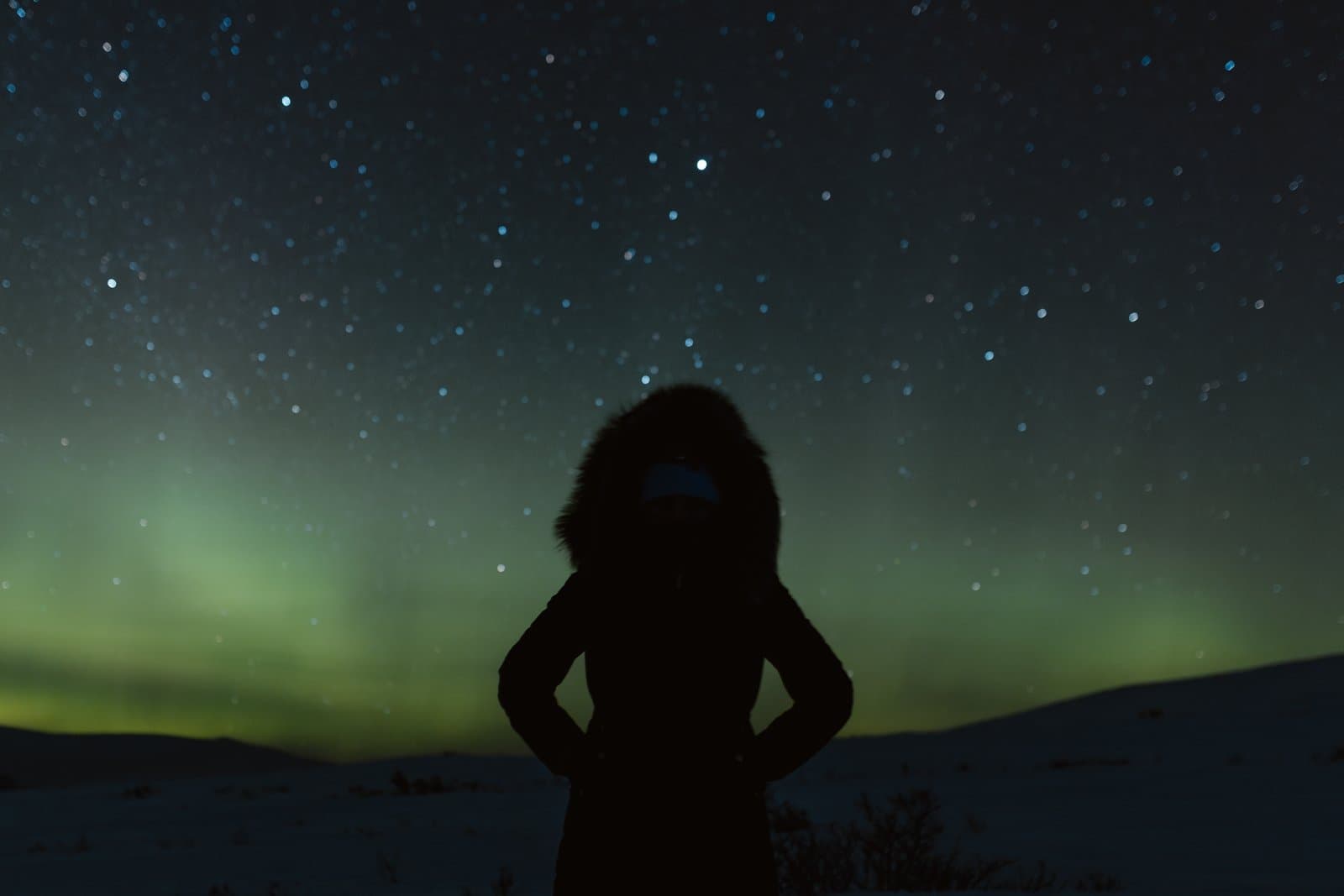 A person in a fur-lined parka stands in a snowy landscape under a vibrant display of northern lights in the night sky.