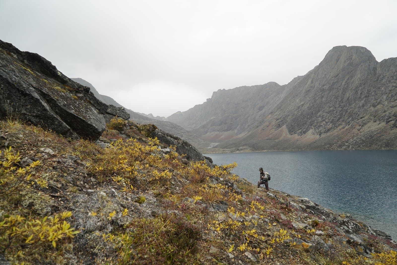 A person wearing a backpack and holding a trekking pole stands on a rocky, vegetated slope with yellow and red fall foliage, overlooking a large blue lake. Rugged, grey mountains rise under a cloudy sky in the background.