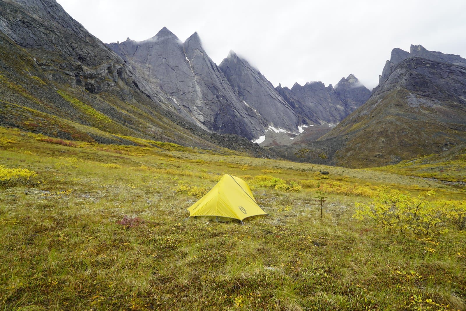 A bright yellow tent is pitched on a vibrant green and yellow valley floor, surrounded by towering, jagged grey mountains under an overcast sky. Patches of snow are visible on the mountain slopes, and the vegetation shows distinct fall colors.