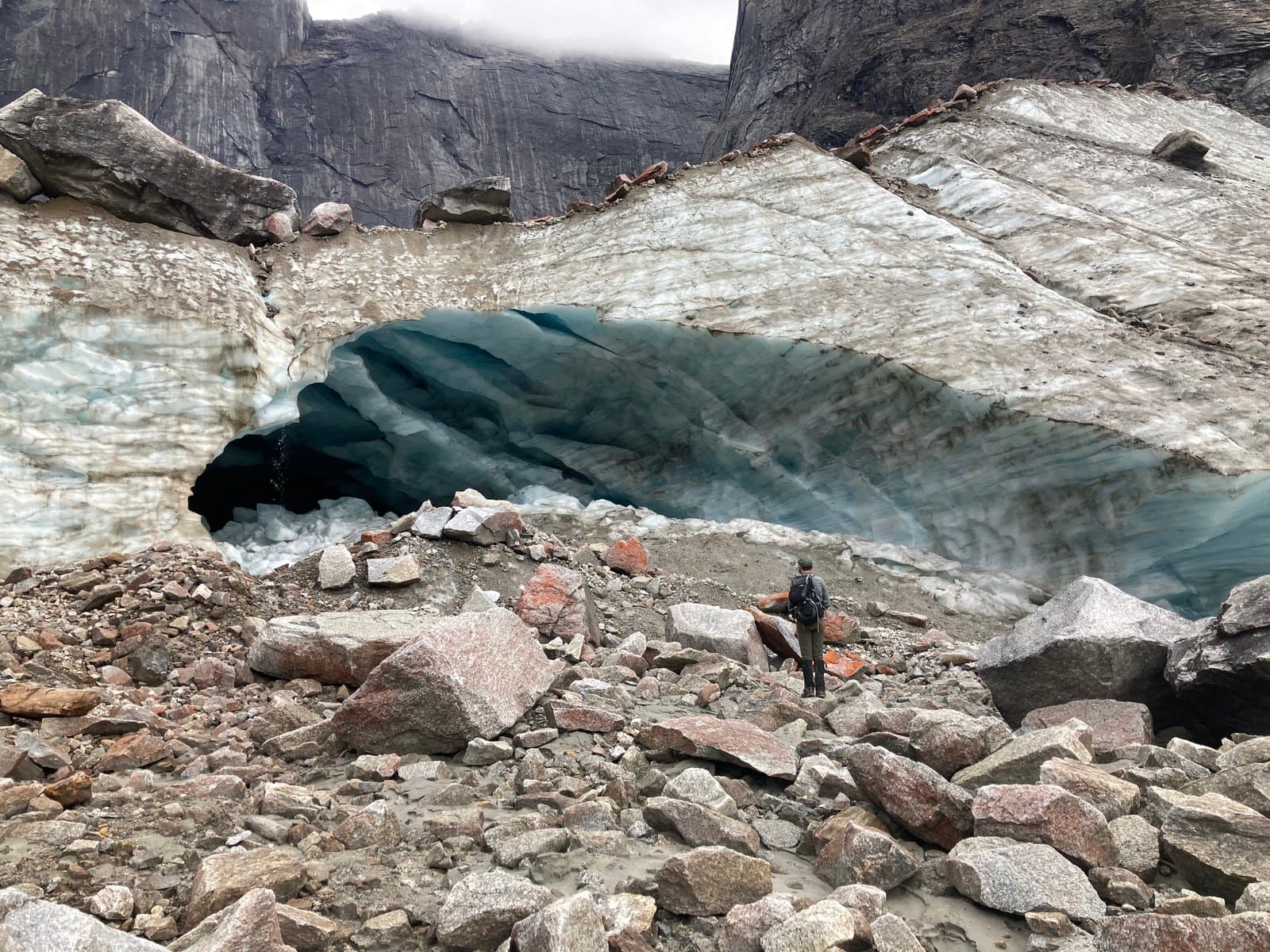 A park ranger, seen from behind, stands in a rocky glacial valley, facing a large, deep blue ice cave entrance. Steep, dark mountain walls rise in the background under an overcast sky.