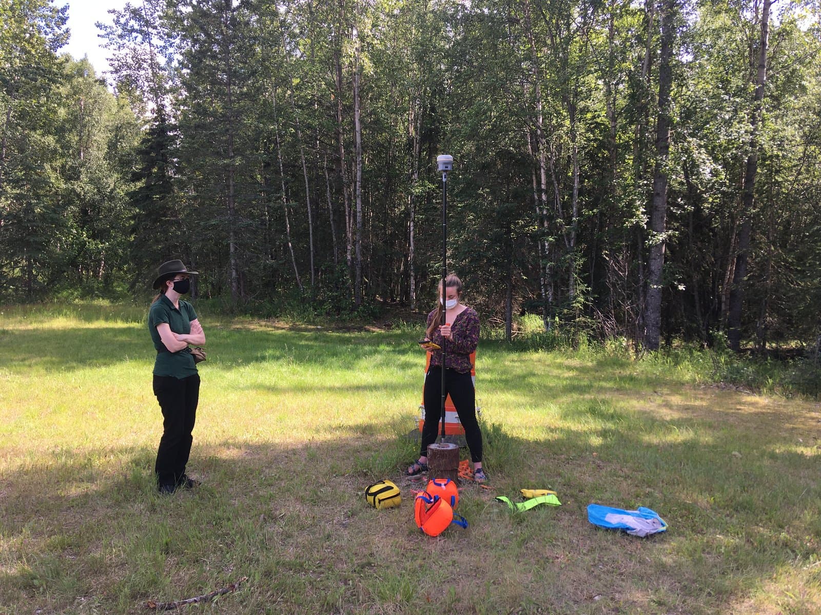 Two women, wearing face masks, are engaged in field work in a sunny forest clearing near Anchorage. One woman stands with arms crossed, observing, while the other operates a surveying instrument on a pole, with various bags of equipment on the ground.