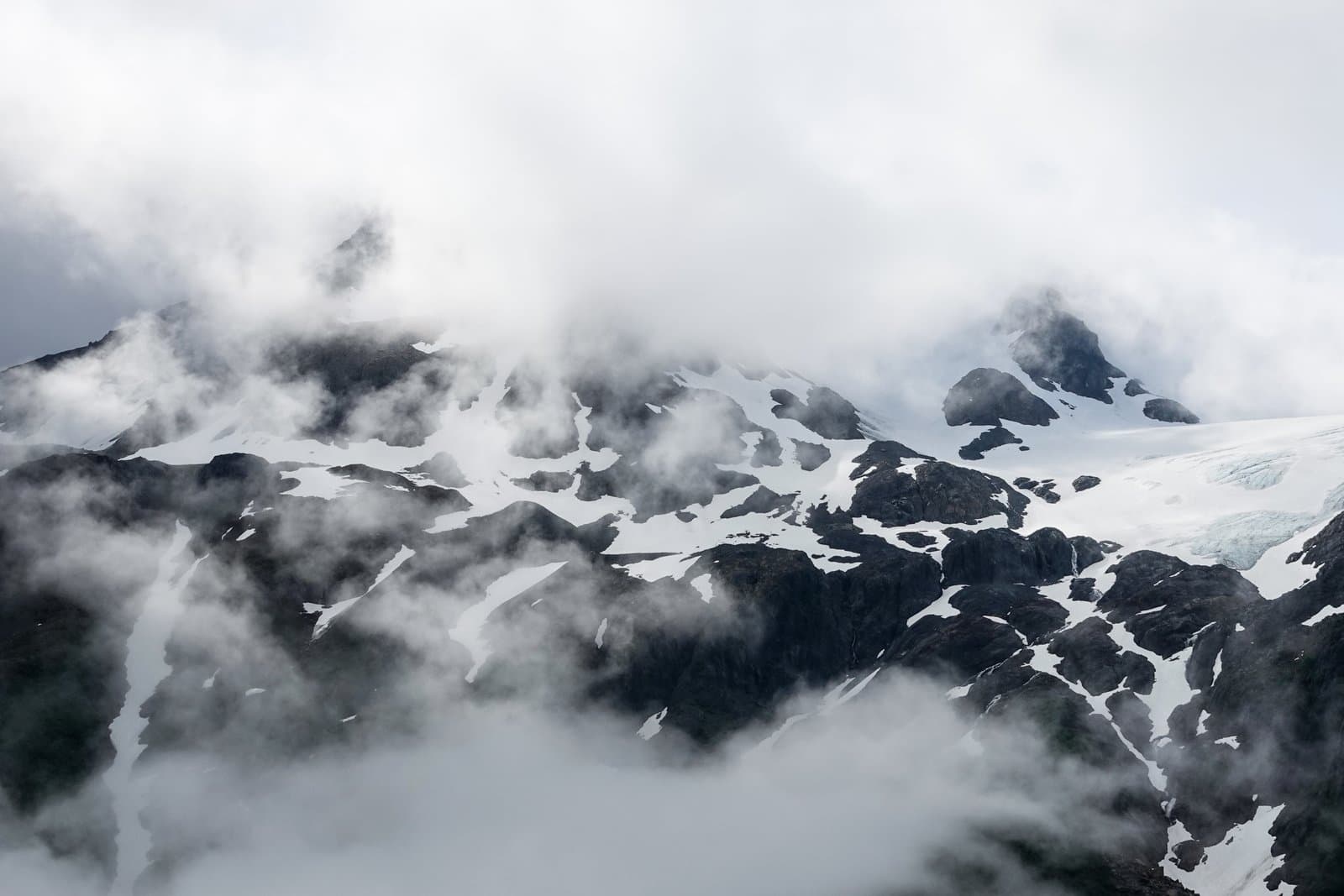 Rugged mountain peaks partially obscured by low-hanging clouds feature patches of snow and glacial ice on a summer day in Alaska.