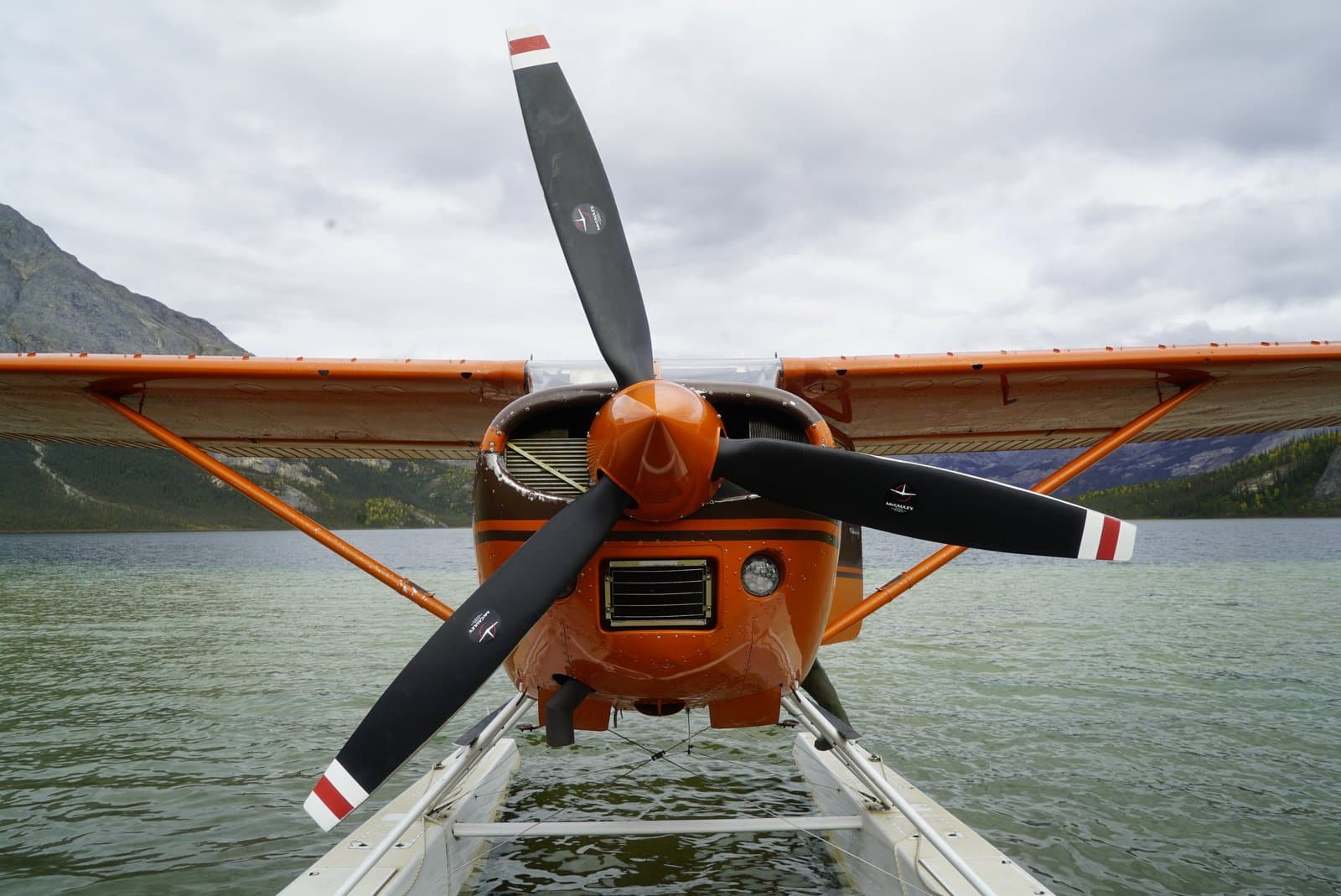 A front-facing view of an orange and brown float plane resting on the calm waters of Takahula Lake, with mountains and forested shores visible under a cloudy sky in the background.