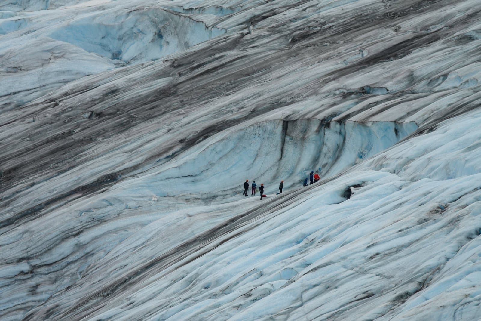 A group of hikers wearing helmets and outdoor gear traverses a crevassed, debris-streaked glacier during a summer excursion.