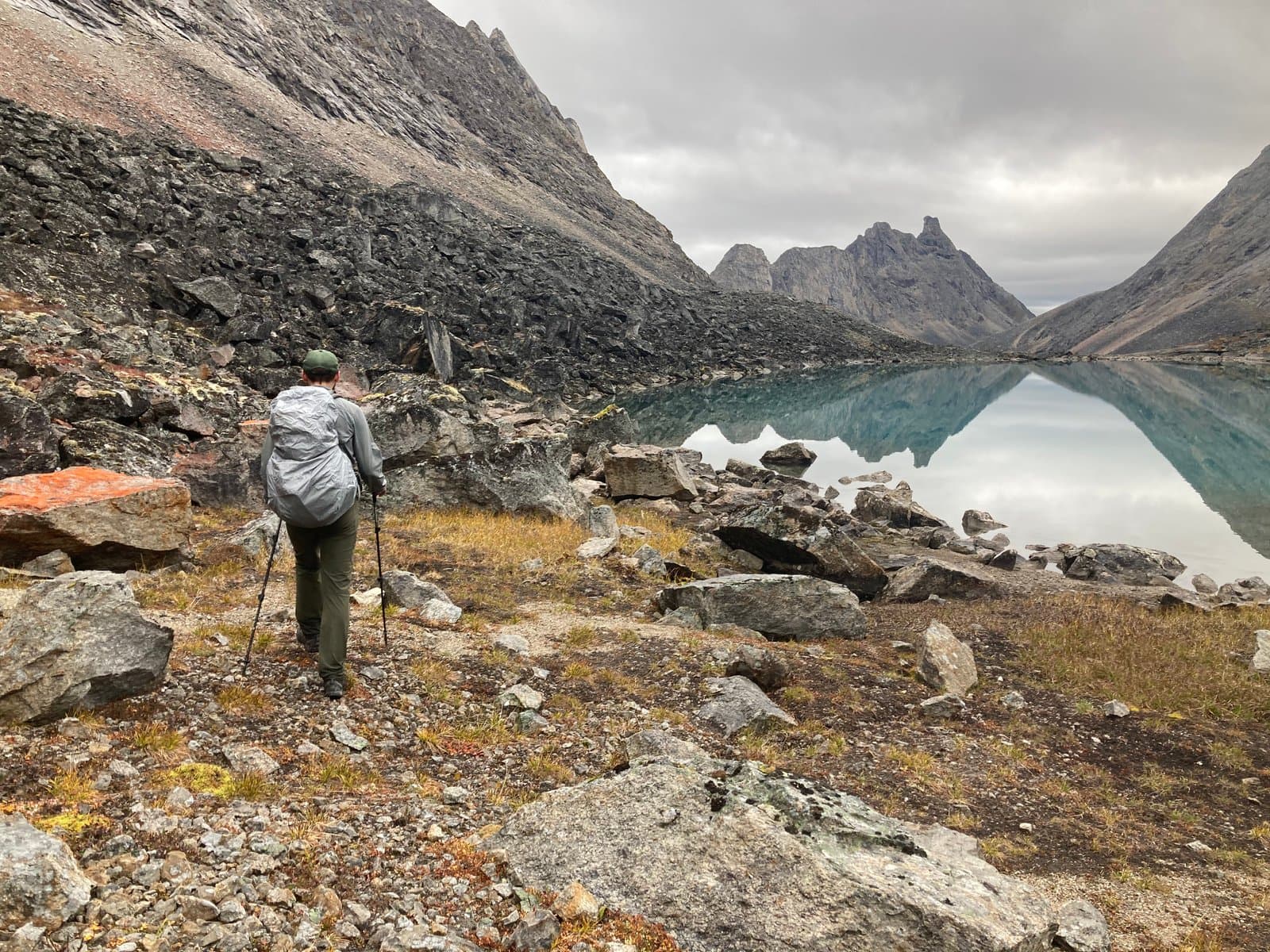 A park ranger, seen from behind with a backpack and trekking poles, hikes along a rocky trail beside a serene, turquoise lake. Rugged, dark mountains dominate the landscape under a cloudy sky, their peaks reflected in the calm water of the lake.