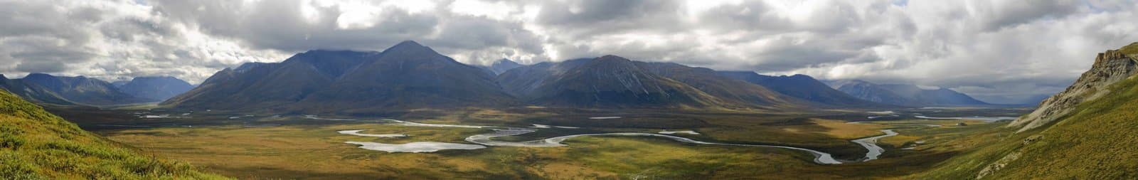 A sweeping panoramic view showcases the vast Noatak River valley in Alaska, featuring a winding river and numerous lakes amidst green and golden tundra. Majestic mountains frame the scene under a dramatic cloudy sky.