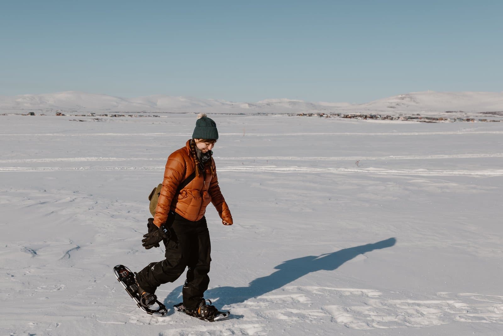 A woman wearing a puffer jacket and beanie walks across a vast, snow-covered landscape on snowshoes under a clear blue sky.