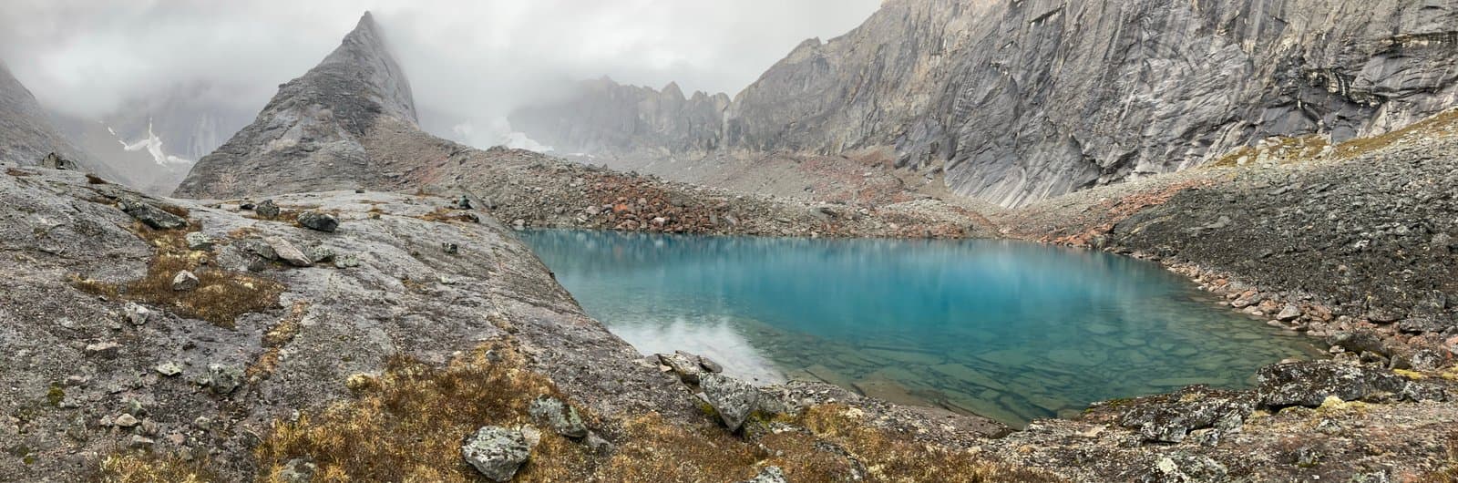 A panoramic view of a vibrant turquoise alpine lake nestled in a rugged, rocky valley. Towering, grey, jagged mountains rise on all sides, partially obscured by low-hanging clouds, with sparse brown and green vegetation on the rocky foreground.
