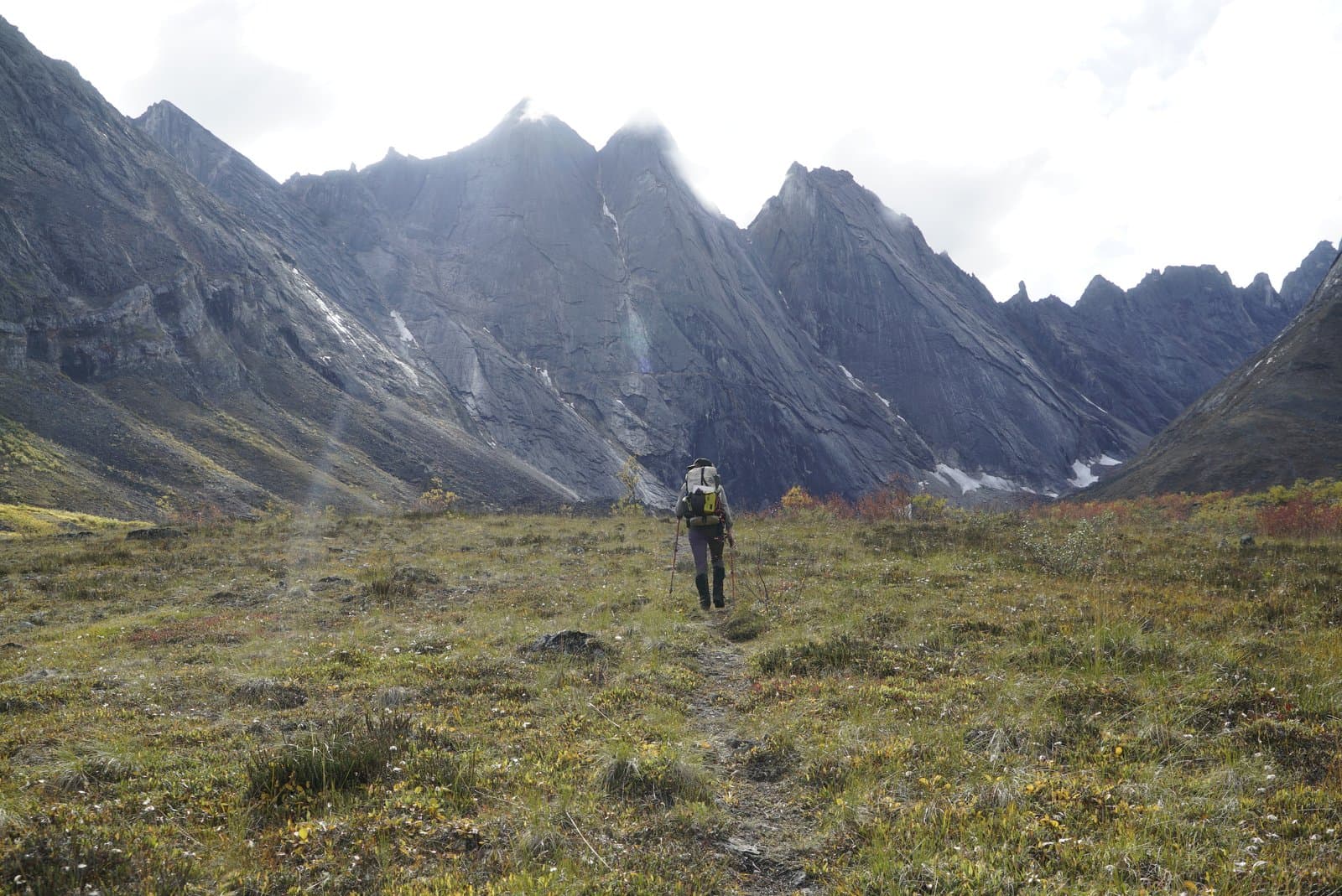 A park ranger with a large backpack and trekking poles walks along a winding dirt path through a vast, tundra-filled valley towards the imposing, jagged Arrigetch Peaks under a bright, cloudy sky. Patches of snow cling to the dark mountain slopes, and the ground vegetation displays early fall colors.