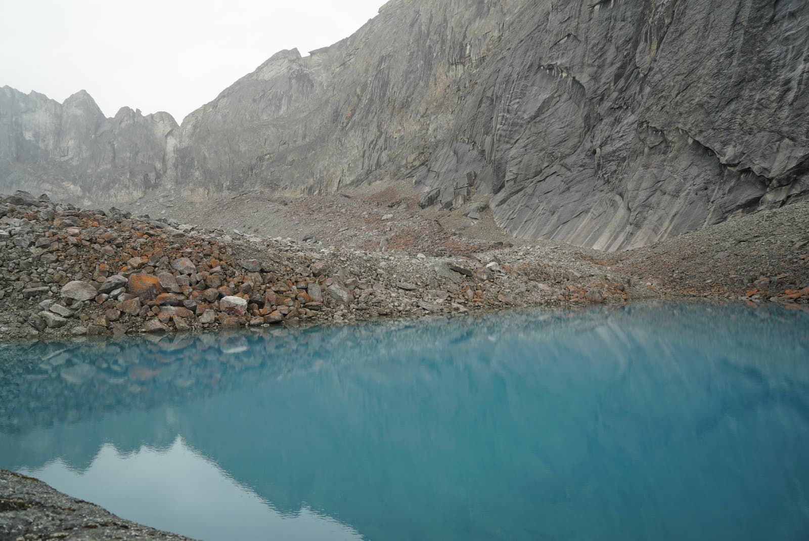 A vibrant turquoise glacial lake is nestled among steep, grey, rocky mountains under an overcast sky. The foreground features a rocky shore with a mix of grey and rust-colored boulders.