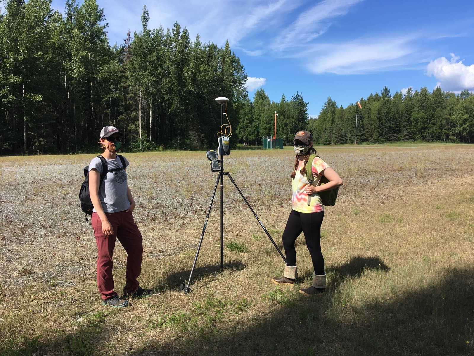 Two women in face masks and outdoor gear stand in a grassy field with a GPS surveying tripod between them, backed by a dense forest and a blue sky with clouds. An orange windsock is visible in the distance, suggesting an airfield.