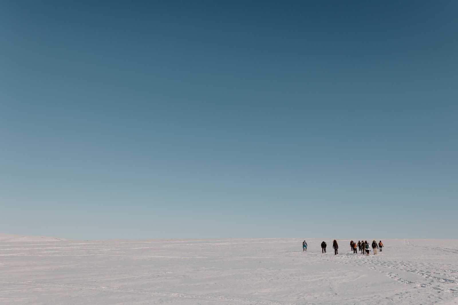 A group of people snowshoe across a vast, flat, snow-covered landscape under a clear blue sky during a winter excursion.