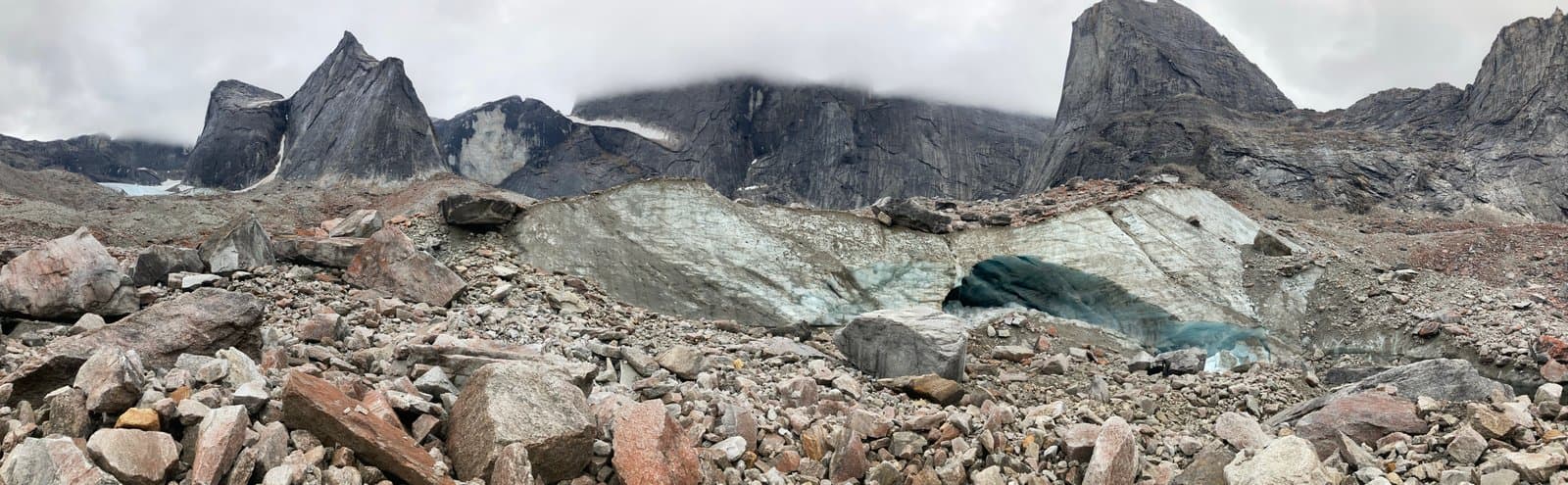 A panoramic view of a rugged mountain landscape in Alaska's Arrigetch Valley, featuring a large glacier with a prominent blue ice cave entrance in the center, surrounded by rocky slopes and towering peaks under an overcast sky.