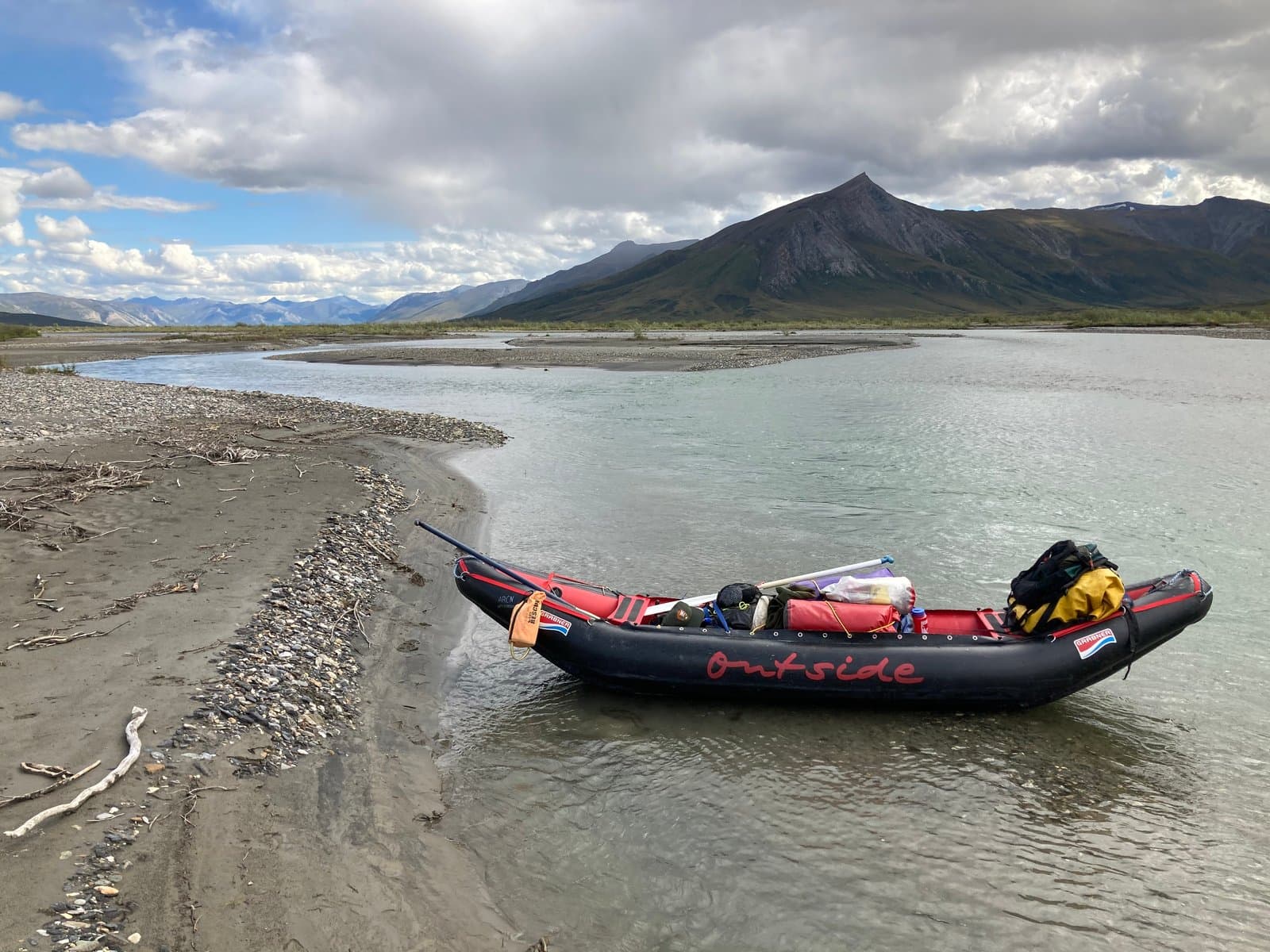 A black and red inflatable raft, loaded with gear, rests partially in the shallow, braided waters of the Noatak River and partially on a sandy, rocky bank. Treeless mountains rise in the background under a cloudy sky.